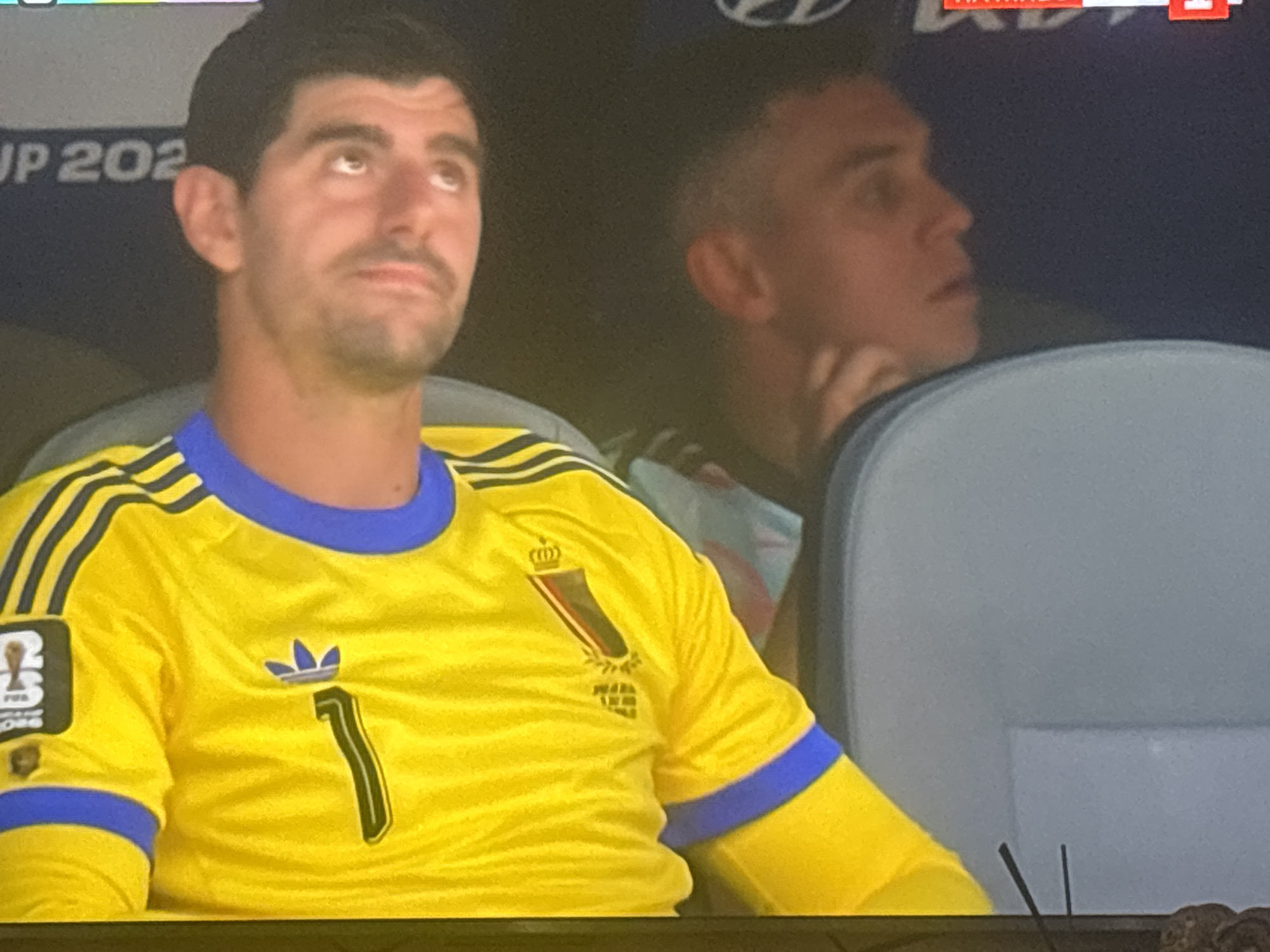 The image shows a close-up of a male soccer player wearing a yellow jersey with blue trim and the number '1' on it. He is looking upwards and to the left. Behind him, another male is visible, also looking to the left. The background is dark, with some text that appears to be from a stadium display, including "JP 202" which suggests it is related to the 2022 FIFA World Cup. The overall setting appears to be a sporting event, likely a soccer match, in an indoor or shaded stadium environment. There is no information in the image to suggest a location in Burgas, Bulgaria.