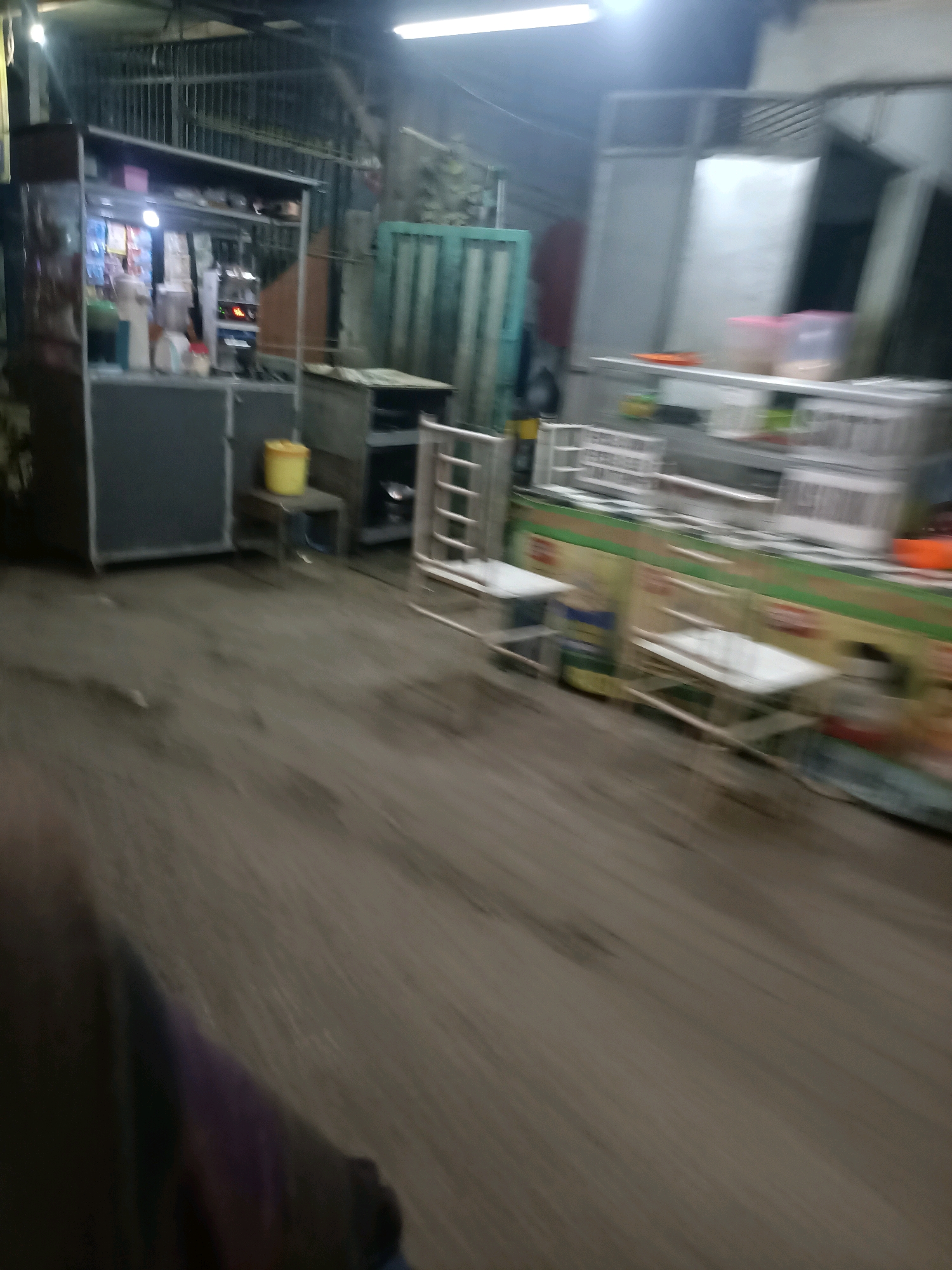 The image shows a blurred outdoor scene, likely a street or a market stall area in Telagaasih, Indonesia. The lighting suggests it is either dusk or night. In the foreground, a blurry person's shoulder and arm are visible, partially obscuring the ground. The ground appears to be unpaved and dusty.
In the middle ground, a food stall or kiosk is visible, with some items displayed on shelves. A yellow bucket sits on a small stool next to the stall. Further to the right, there are several metal shelving units and what appear to be plastic containers or boxes. There are also chairs and tables, suggesting a seating area for customers. The background shows a building with corrugated metal siding and a metal fence. The overall impression is of a modest, possibly temporary, commercial establishment. The blurriness of the image makes it difficult to identify specific items or people with clarity.