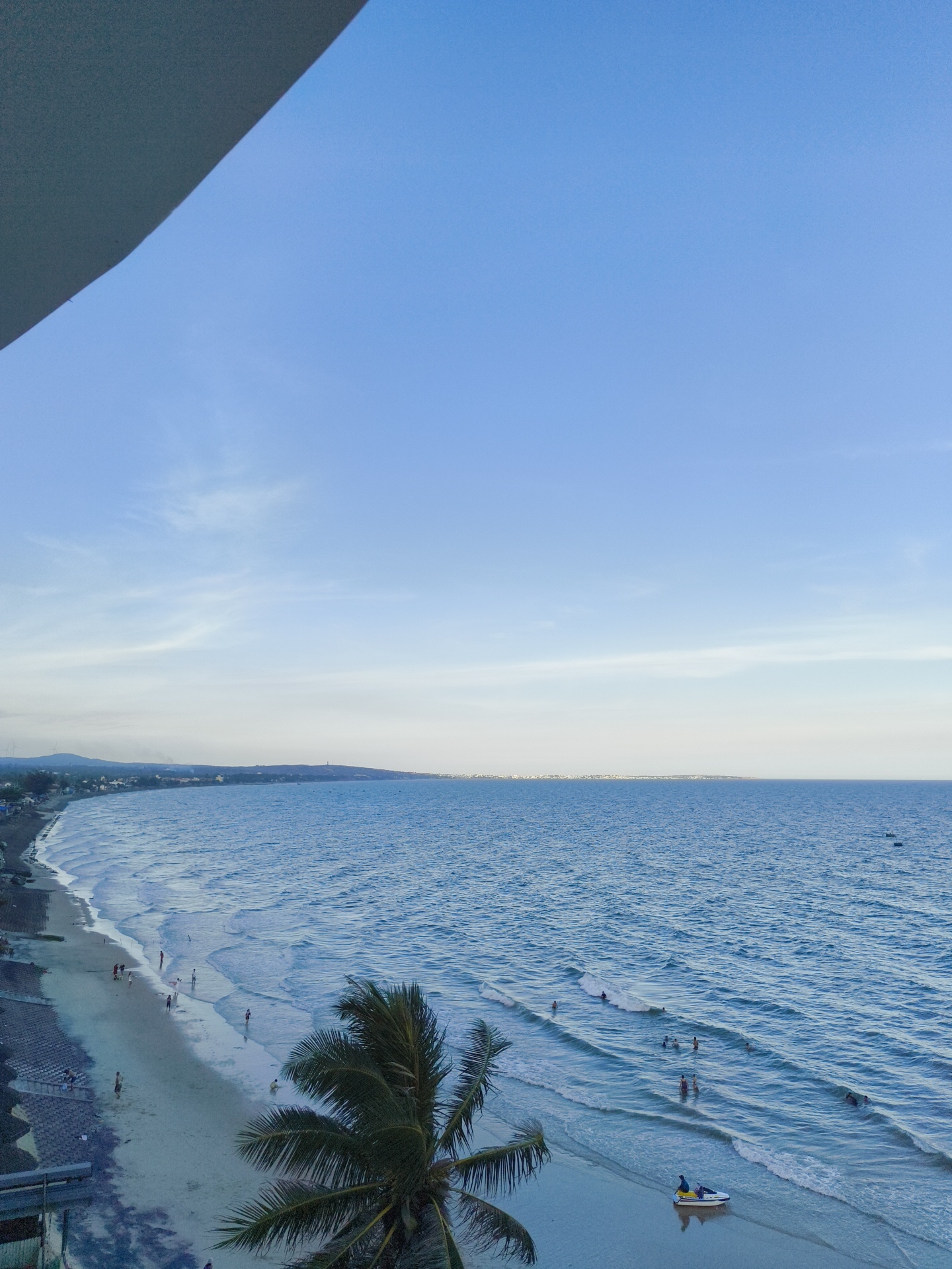 This is an aerial view of a beach in Phu Thuy Ward, Vietnam. The scene captures a wide expanse of the ocean with gentle waves lapping at the shore. The beach is sandy and appears to be populated with several individuals engaged in various activities. Some people are seen walking along the shoreline, while others are wading in the shallow water. In the distance, a jet ski is being operated by one person, and there are a few other individuals in the water near it, suggesting recreational use of the sea. A prominent palm tree with lush green fronds is visible in the foreground, partially obscuring the view of the beach. The coastline stretches into the distance, with a hint of a settlement or town visible along the horizon, characterized by scattered buildings. The sky is a clear blue with a few wispy clouds, indicative of fair weather. The overall impression is a serene coastal environment, likely during daylight hours.