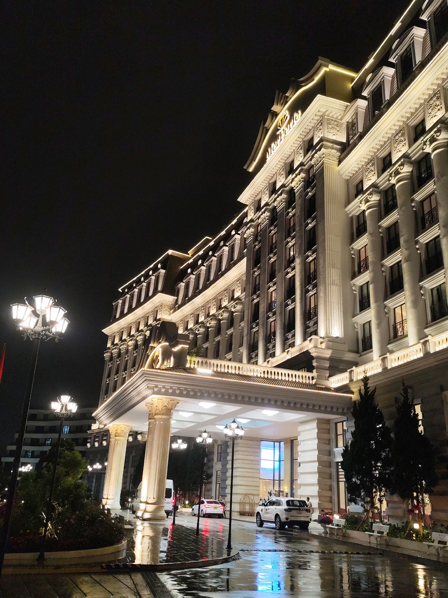 The image displays a grand, multi-story hotel building at night, likely located in Da Lat, Vietnam. The hotel, identified by the partially visible "Mer Perle" sign on its facade, exhibits an ornate, classical architectural style with numerous columns, balustrades, and intricate decorative moldings. The building is brightly illuminated with warm, golden-toned exterior lighting, highlighting its architectural features against the dark sky.
The scene centers around the hotel's entrance, featuring a large, covered porte-cochère supported by prominent columns. The ground, including the driveway and surrounding pavement, is visibly wet and reflective, indicating recent rain or damp conditions. This wet surface creates shimmering reflections of the hotel's lights, street lamps, and vehicle headlights and taillights.
Several vehicles are present at the entrance: a white SUV is positioned under the covered area, and a white sedan and a white van are also visible on the wet roadway. A few indistinct human figures can be observed near the vehicles and the entrance, suggesting activity like guest arrivals or departures. Tall, multi-globed street lamps provide additional illumination, complementing the hotel's extensive lighting. Landscaped areas with small trees and bushes are visible in the foreground.
The time of day is night, and the weather is damp. The overall impression is one of luxury and elegance, captured in the dramatic interplay of light and reflections.