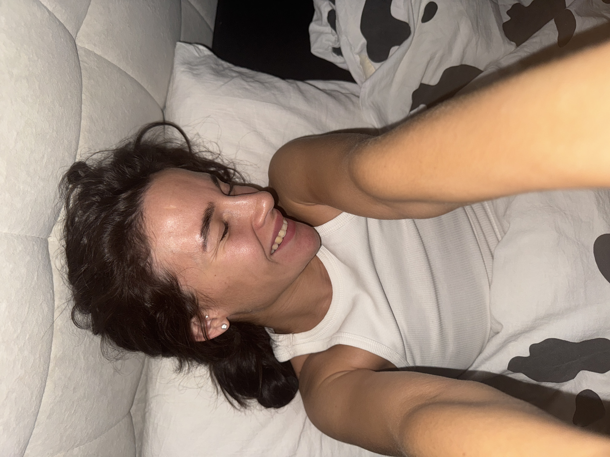 A woman is lying in bed, smiling. She has dark hair and is wearing a white tank top. The bed has a white headboard and white sheets, with a cow-print blanket visible. The location is not discernible from the image.