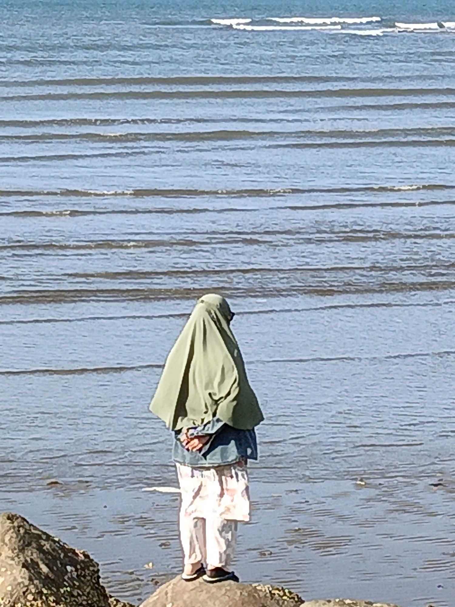 A person wearing a headscarf and loose-fitting light-colored pants stands facing the ocean on a rocky shoreline in Bulukumba, Indonesia. The person's hands are clasped behind their back, and they are wearing a green headscarf and a blue denim jacket over a white and pink patterned top. The ocean has gentle waves, and the sky is a clear blue. The foreground shows a large rock with texture.