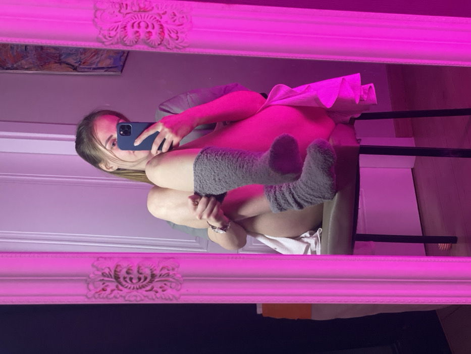A mirror selfie captures a blonde-haired woman with a phone in hand, seated in a chair. She wears gray, knee-high socks. A white towel is partially visible under her. The reflection showcases the ornate frame of the mirror, along with a glimpse of artwork on the wall behind her. The image is heavily illuminated with pink light.