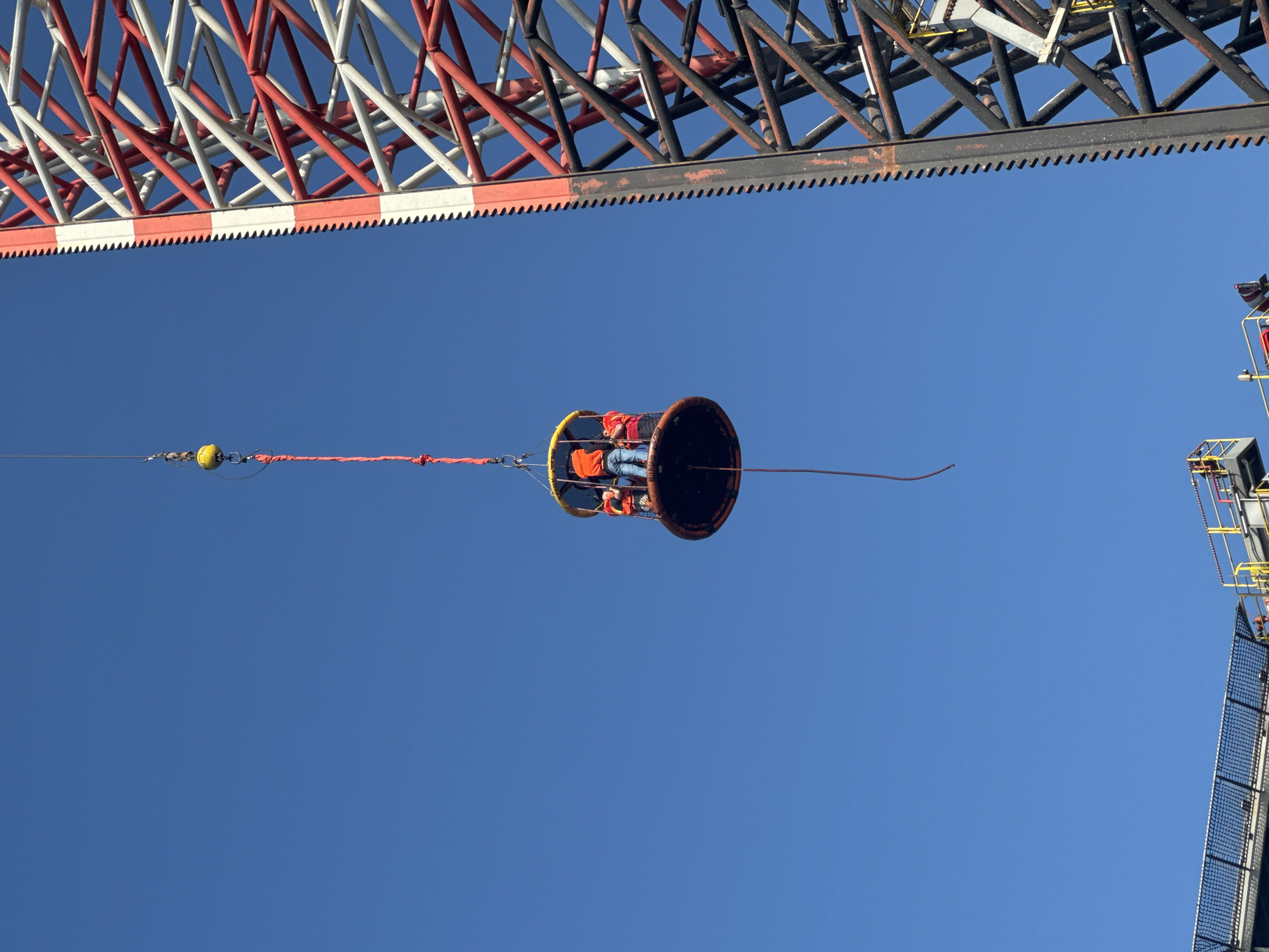 In an outdoor, clear blue sky, a large industrial red and white lattice tower dominates the right side of the frame. Hanging from a red rope attached to a yellow buoy, a circular open-air basket carries three people. Two individuals are visible from the back, wearing orange high-visibility vests. One person in the basket appears to be wearing blue jeans and an orange top. A dark rope hangs down from the basket. In the lower left portion of the image, a section of a lower structure with a black netted surface and yellow railings is visible, suggesting a platform or helideck. The scene appears to be an industrial or offshore environment, likely involving maintenance or transport at height. The exact location in the city of Unknown, Italy cannot be determined from the visual information provided.