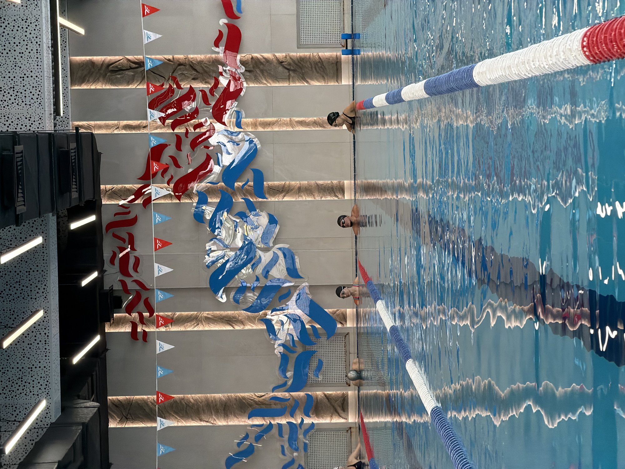 An indoor swimming pool in Bishkek, Kyrgyzstan features swimming lanes marked by blue and white ropes. Several individuals are visible in the water, some wearing swim caps and goggles. The pool area is decorated with red and blue wave-like art installations on the wall, and a string of triangular flags in red, white, and blue hangs above. The lighting appears to be modern, with long fluorescent fixtures on the ceiling. The reflections of the lane ropes and people are visible on the surface of the water.