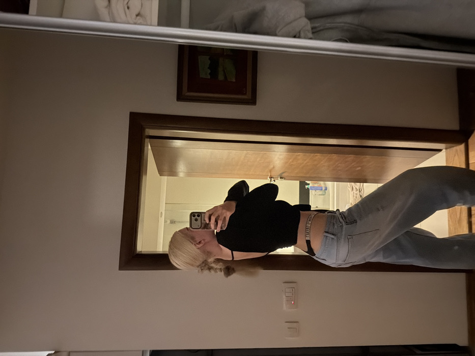 The image displays an interior scene depicting a female individual taking a mirror selfie. The subject, with blonde hair styled in two braids, is positioned in profile, looking over her left shoulder towards a smartphone held in her right hand. She is wearing a black long-sleeved, open-back top and light-wash blue jeans. A "Victoria's Secret" branded waistband is visible above the jeans, crossing her lower back.
The environment appears to be a room within a dwelling. A large full-length mirror occupies the right side of the frame, reflecting the subject and parts of the room. Behind the subject is a plain, light-colored wall. To the subject's right, a wooden doorway opens into an adjacent room, identifiable as a bathroom, where a white bathtub/shower fixture and several bottles on a ledge are visible. On the wall to the left of the doorway, two light switches are mounted. To the right of the doorway (within the mirror's reflection), a framed piece of art hangs on the wall. The scene is illuminated by artificial indoor lighting. This interior scene is stated to be located in Budva, Montenegro, though no specific geographical markers within the city are visually identifiable from the image itself.