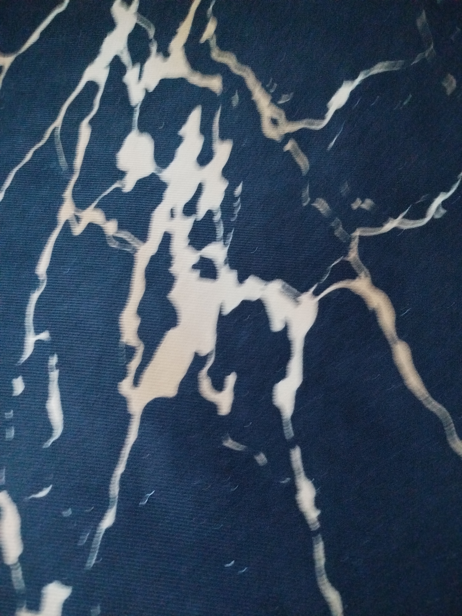 The image displays a close-up, abstract view of a dark blue surface with irregular, lighter-colored veining, resembling marble or a similar stone pattern. The veining is predominantly off-white and light tan, with some areas appearing brighter than others, creating a sense of depth and texture. The pattern is organic and flowing, with thin, branching lines and broader, irregular patches. The dark blue background has a subtle, woven texture, possibly indicating fabric or a similar material. There are no discernible objects, people, or environmental features visible in the image, nor is there any information about its location in the city of Unknown, Unknown.