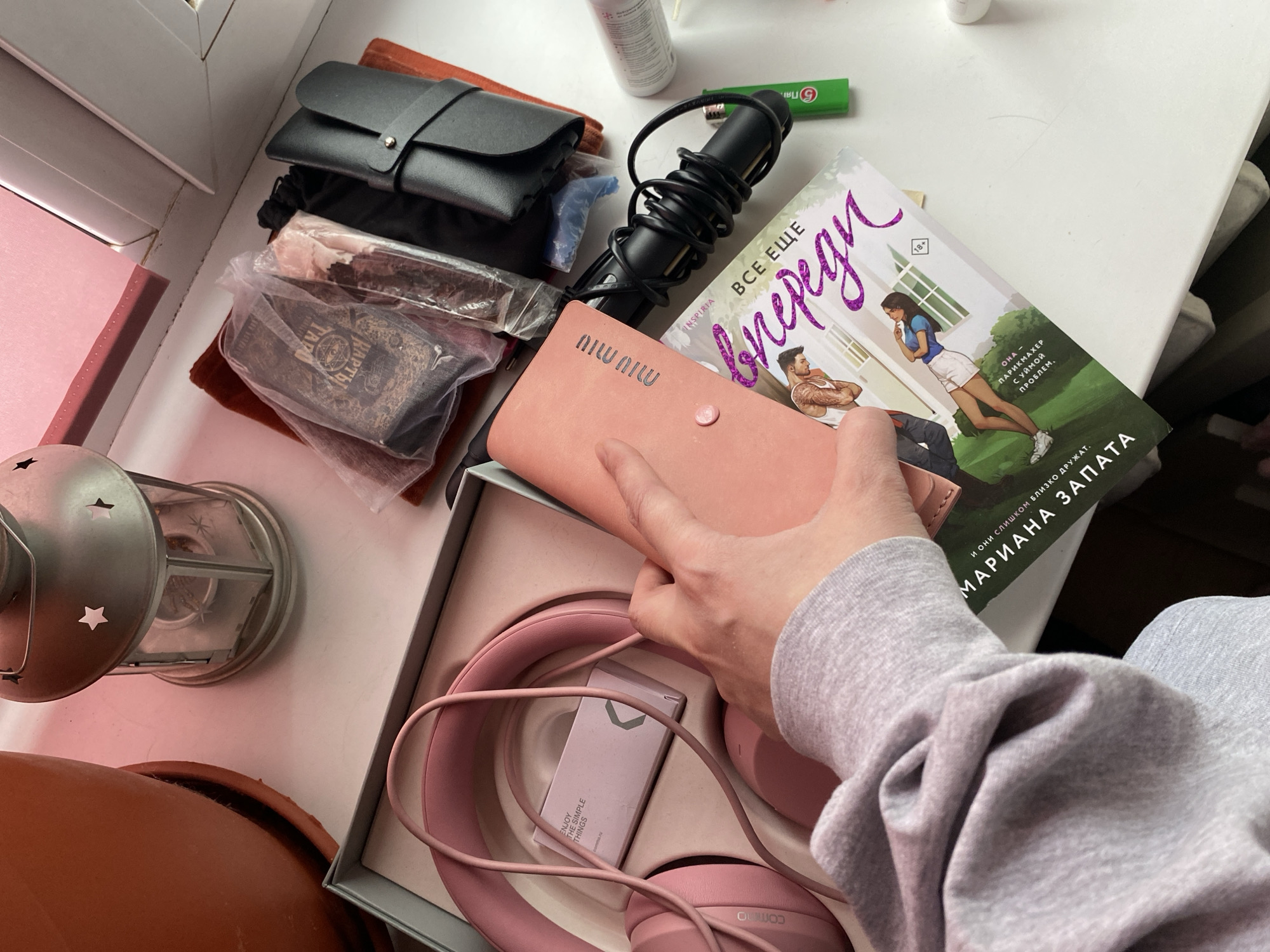 A high-angle shot captures a cluttered scene, likely on a table or surface near a window. The composition includes an assortment of personal items: a black leather glasses case, a deck of tarot cards wrapped in plastic, a black curling iron with a coiled cord, a pink Miu Miu cardholder being held by a hand, a book with a cover featuring a man and a woman, and a box containing pink Commute headphones. Other items include a green pen, a white spray bottle, a silver lantern, and an orange clay pot. The hand holding the cardholder belongs to someone wearing a grey sweatshirt. The book appears to be in Russian, with the title in large letters, "ВСЕ ЕЩЕ Впереди". There is no evidence to suggest that the location is in Votkinsk, Russia.