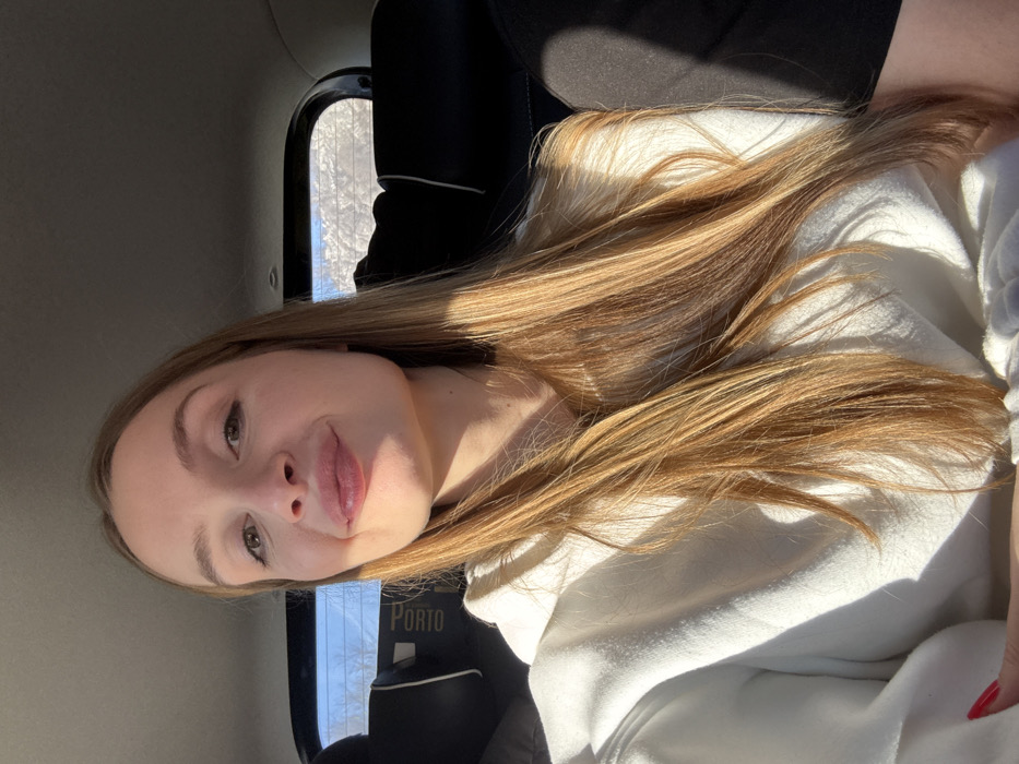 A young adult female is depicted in a self-portrait, seated in the passenger compartment of a vehicle in Stubica, Montenegro. She has long, straight blonde-brown hair, a light complexion, and is looking directly at the camera with slightly pursed lips. She is wearing a white hooded sweatshirt, and red nail polish is visible on her left hand. Strong natural light illuminates her right side, creating shadows. In the background, the vehicle's dark interior headrests are visible, with one to the left of the subject marked "PORTO" vertically in white text. A blurred exterior scene through a window shows a bright blue sky above a white, possibly snow-covered, landscape.