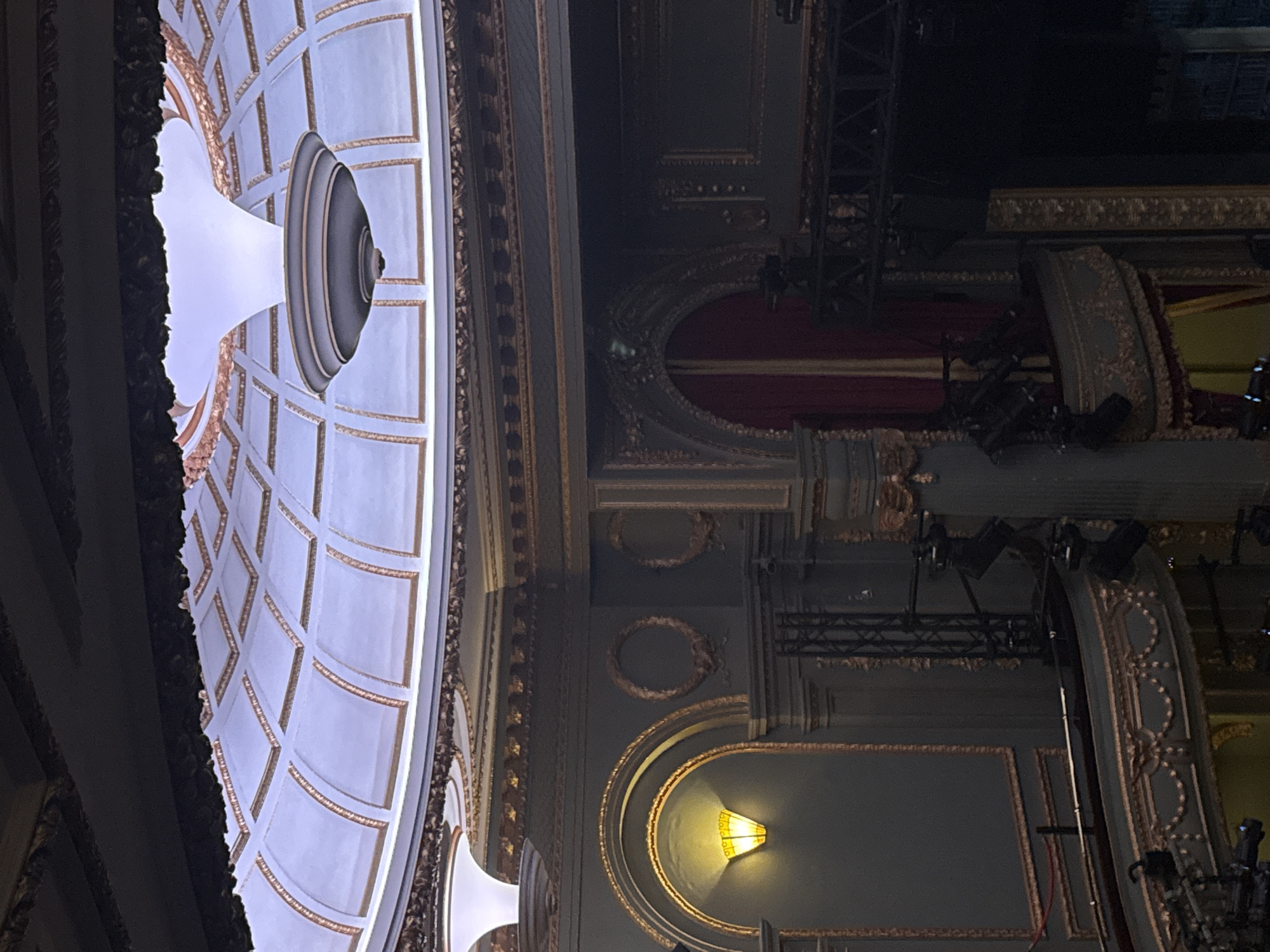 The image shows the interior of a theater. The ceiling has a dome-shaped design with a central lighting fixture. The walls are ornate with architectural details including arched structures, decorative moldings, and wall sconces. Stage lighting equipment is visible in the upper right portion of the image. The theater's architectural style appears to be classical or Baroque. The location in London is inferred based on the theater's architectural style commonly found in London's historic theaters.