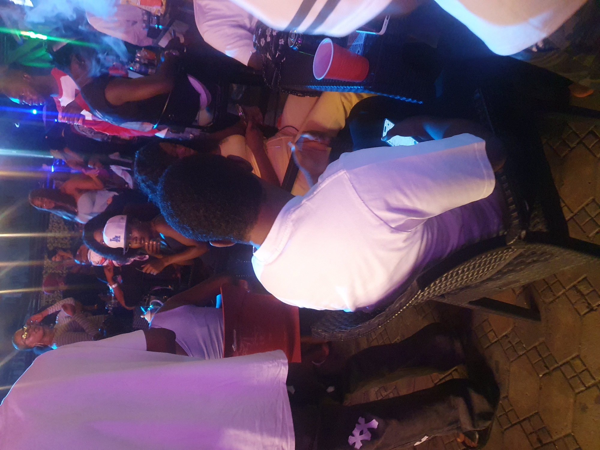 The image depicts a dimly lit indoor setting with multiple people gathered, possibly at a nightlife venue in Lokoja, Nigeria. A group of individuals is seen seated around tables, several with beverages such as red plastic cups. Some are using their phones or engaging in conversation. Some figures are more clearly visible, including one person wearing an LA cap and another wearing a white shirt. The environment is further suggested by the tiled floor. The lighting is provided by some electric lights from the side. The presence of people, beverages, phones, and a tiled floor indicates the potential venue.