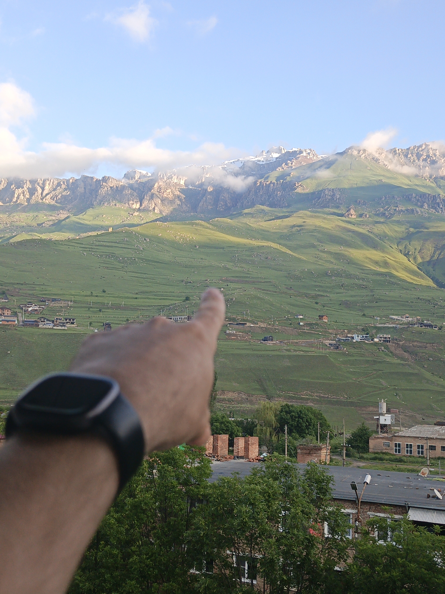 An adult's left arm and hand with a smartwatch are in the foreground, obscuring the lower left portion of the image. The hand is pointing towards a distant, green, mountainous landscape. In the middle ground, there is a village with various buildings, including houses, a water tower, and what appears to be an industrial facility or storage area with brick structures. The background features a large, imposing mountain range with rocky peaks, some of which are capped with snow and partially obscured by clouds. The sky is a light blue with scattered white clouds. The scene appears to be taken during daylight. The geographical location is the village of Verkhny Fiagdon, Russia.