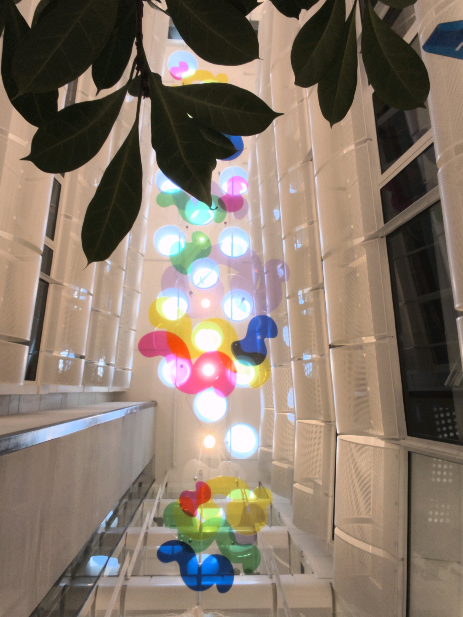The image shows an interior view looking upwards, featuring a tall installation of colorful, translucent geometric shapes that appear to be made of plastic or acrylic. These shapes, in shades of blue, yellow, pink, and green, are lit from within, creating a glowing effect. They are suspended from above by thin wires. Overhanging the top of the scene are large, dark green leaves of a plant, possibly a tree branch. The background consists of a modern architectural structure with vertical white panels, some of which have a perforated or textured surface, and dark-framed windows. A light-colored staircase with a metal railing is visible on the left side. The overall impression is of an art installation within a contemporary building. This scene is located in the interior of the Tallink City Hotel in Tallinn, Estonia, where a similar multi-story sculpture of colored acrylic forms is featured in the atrium. There are no people visible in the image.