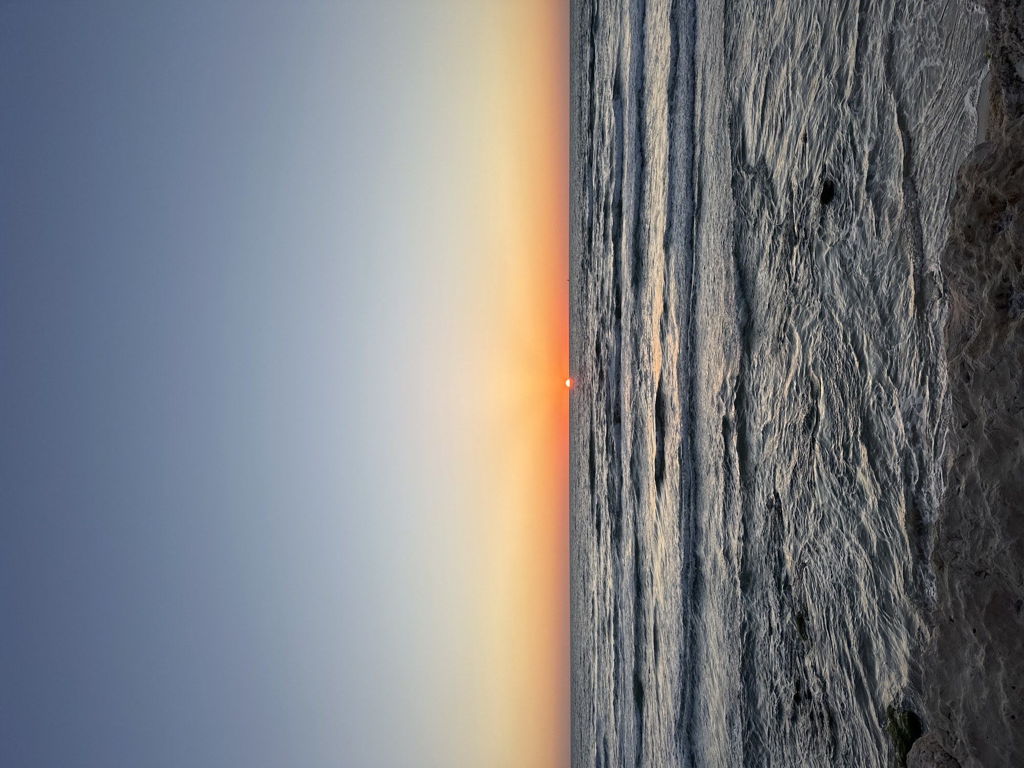 The image, oriented vertically, depicts a sunset over a body of water with a coastline on the right, specifically identifiable as Tel-Aviv, Israel. The sky, occupying the upper half of the frame (when viewed correctly in landscape orientation), displays a gradient of colors: a deep slate-blue on the far left transitions to warm oranges and yellows around the setting sun, then fades to a soft, hazy blue-grey on the right.
The sun, a distinct, circular orange-red orb, is positioned low on the horizon, creating a bright focal point. Below the crisp horizon line, the ocean fills the lower half of the frame. It is characterized by numerous parallel wave patterns, their textured surfaces reflecting the fading light of the sun. The waves appear to be moving towards the shore or viewer. On the far right edge of the image, a darker, textured area suggests a rugged coastline, possibly a rocky beach or cliffside, contrasting with the rhythmic patterns of the water.
The scene captures the tranquil moment of dusk with clear weather conditions. No people or visible text are present. The overall impression is one of natural beauty and serenity as day transitions to night.