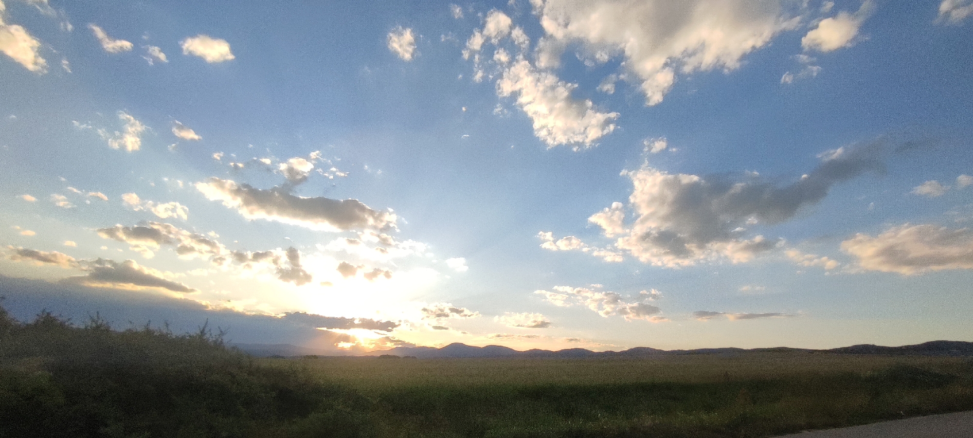 This is a wide-angle, eye-level, outdoor photograph taken in Momin Prohod, Bulgaria. The scene depicts a rural landscape at sunset. The sky is a clear blue, scattered with fluffy white and gray clouds, some of which are backlit by the setting sun. The sun itself is low on the horizon, peeking through a band of clouds, casting a warm, golden light across the scene. In the foreground, there is a field of green grass and some dark green foliage, possibly bushes or trees, lining the left edge of the frame. In the distance, a range of low, dark mountains forms the horizon line. The overall impression is one of a serene and tranquil natural setting. There are no people, animals, or man-made structures immediately visible, other than what might be a faint road on the lower right edge.