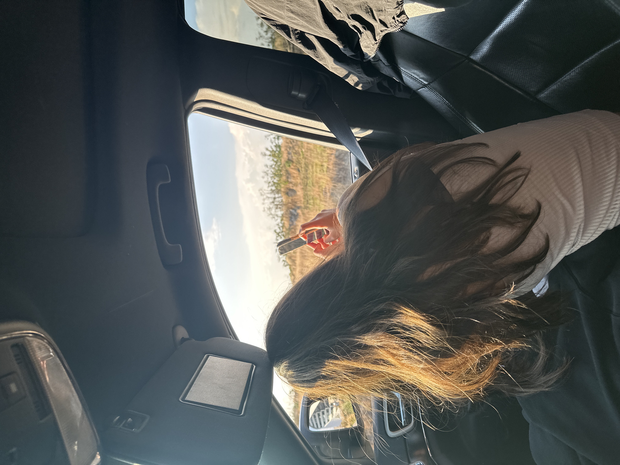 A person is leaning out of the window of a dark-colored car, holding a mobile phone in their hand. The person has long, dark hair with sunlit ends and is wearing a white ribbed top. Part of the car's interior is visible, including the visor, roof, leather seats, and a seatbelt. Outside the window, there's a view of a landscape with dry vegetation under a bright, clear sky. Based on the prompt, this is in the city of San Bernardino Tlaxcalancingo, Mexico.