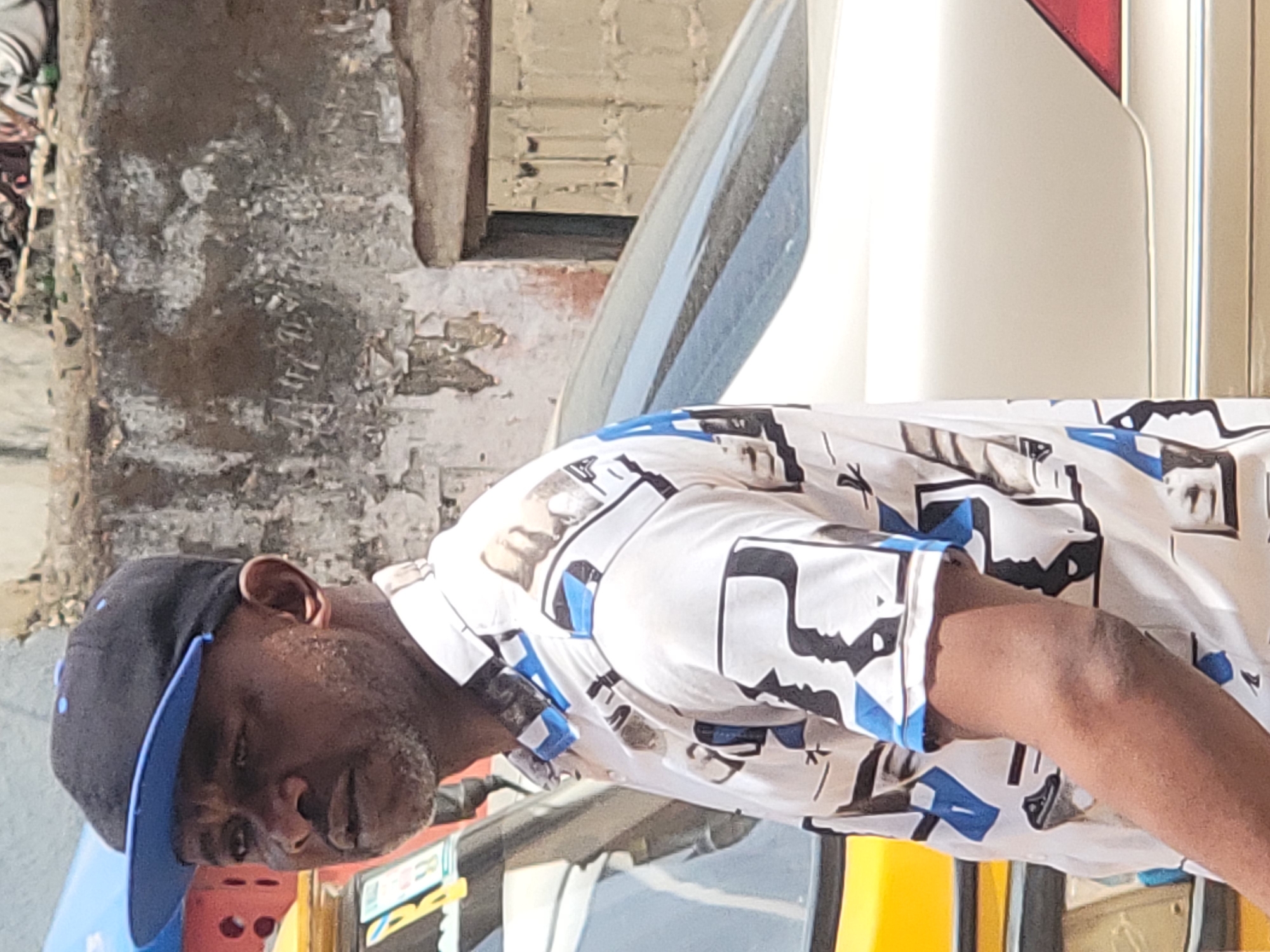 A man wearing a black and blue baseball cap and a white shirt with a geometric print stands outdoors in Maiburiji, Nigeria. He is positioned near a vehicle, with weathered buildings in the background.
