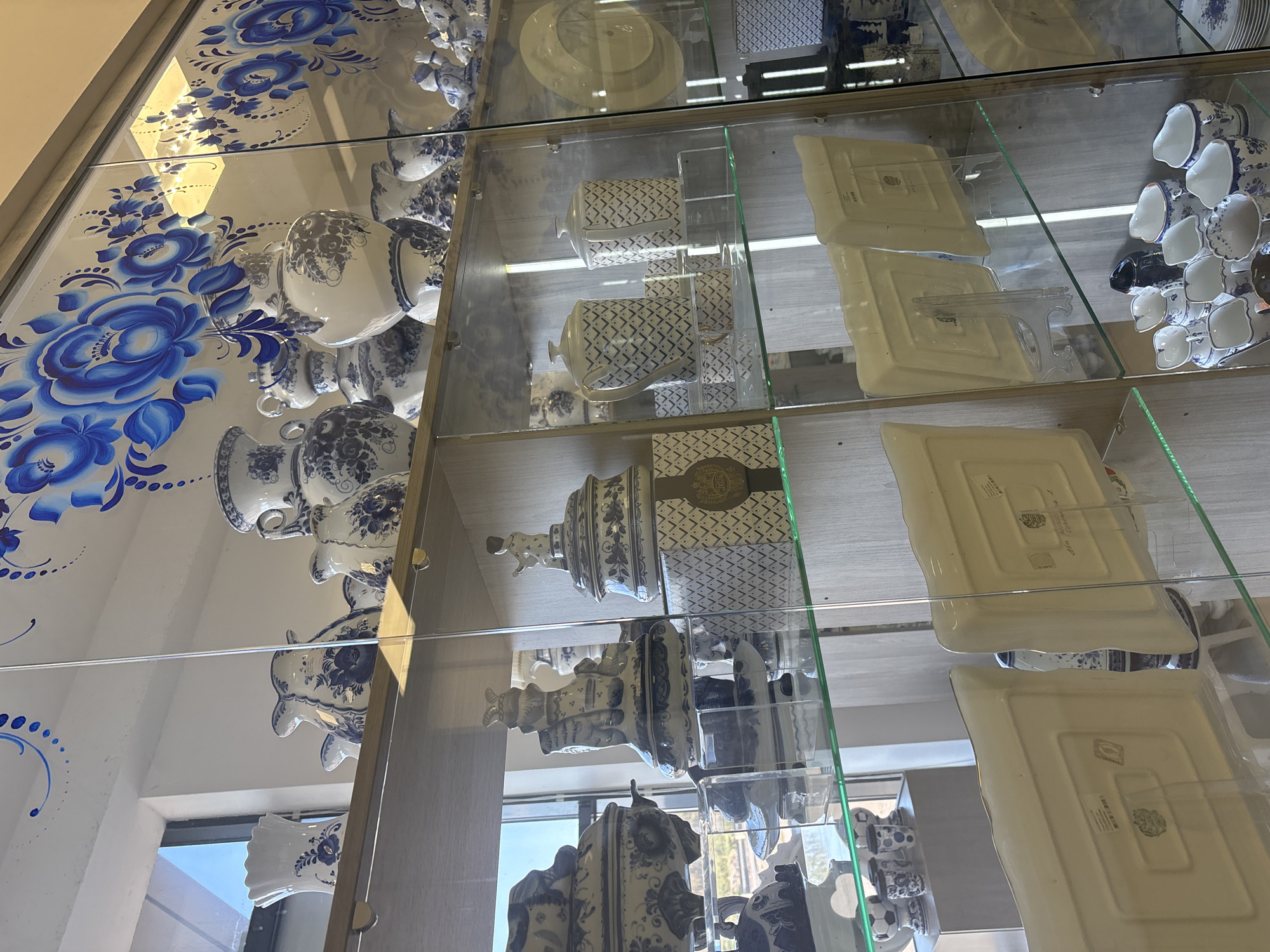 The image shows a display cabinet filled with blue and white ceramic dishes and figurines, likely Gzhel porcelain. The cabinet appears to be inside a building in Речицы, Russia, based on the Russian Gzhel style. The display includes plates, cups, teapots, and figurines with floral patterns. The cabinet is made of glass and metal, and the items are arranged on multiple shelves. Natural light is entering through a window, creating reflections on the glass. The walls have a decorative blue floral pattern.