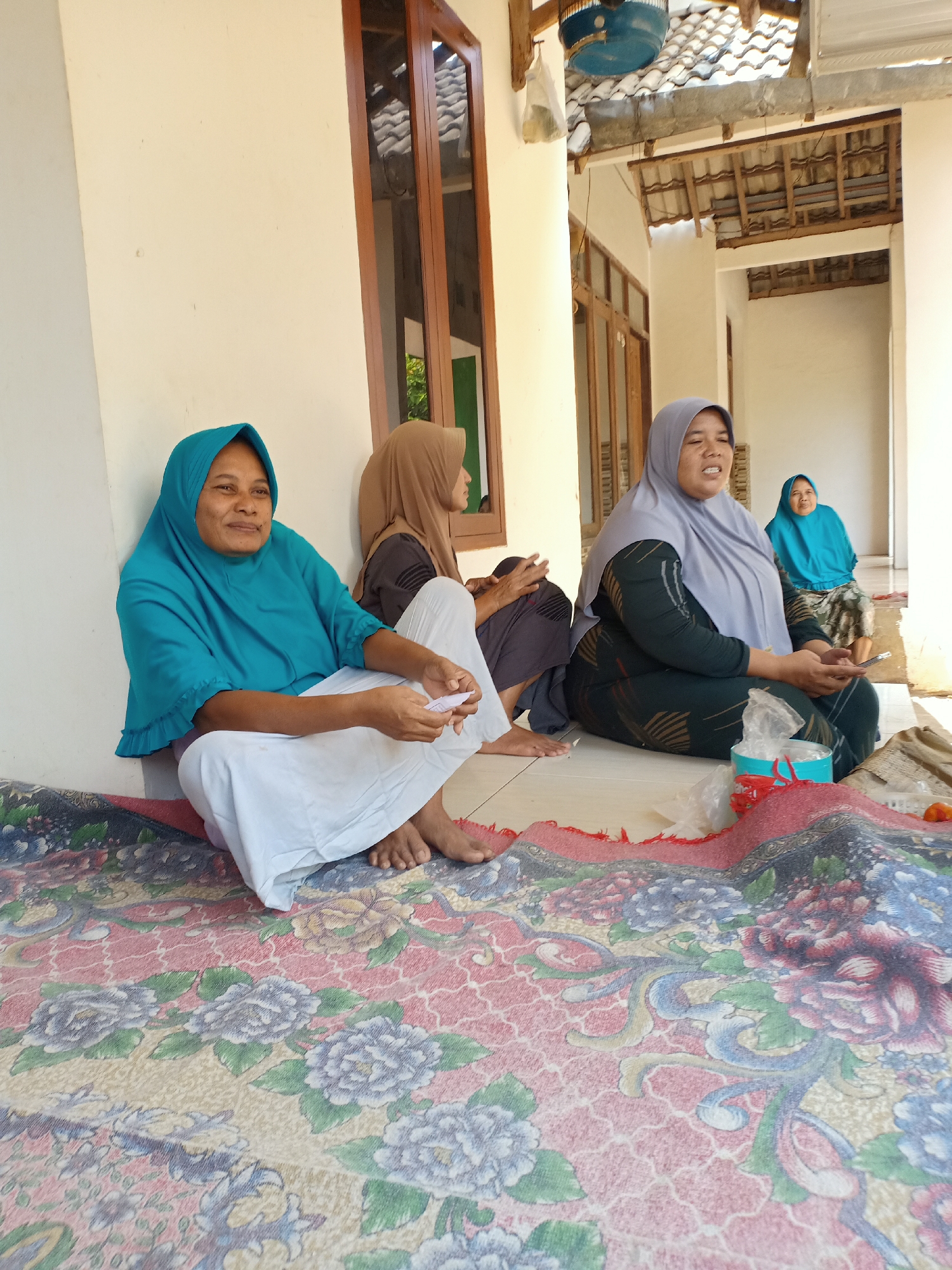 Four women are sitting on the ground in front of a building in Palang, Indonesia. The woman on the far left is wearing a turquoise hijab and white pants. She is holding a piece of paper in her right hand. The woman next to her is wearing a brown hijab and dark pants, and she is looking at her phone. The woman in the center is wearing a grey hijab and a dark patterned dress. She is speaking and looking to her left. The woman on the far right is wearing a blue hijab and a patterned skirt. She is looking to her left. The women are sitting on a colorful rug with floral patterns. The building behind them appears to be made of white stucco with wooden window frames. There is a tiled roof visible above the building. The scene is outdoors and appears to be daytime, with sunlight casting shadows.