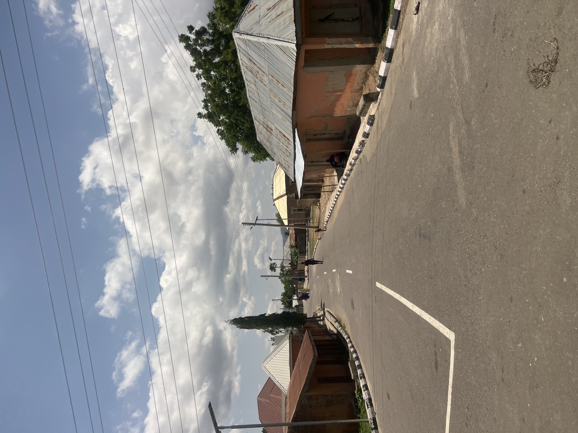 The image presents a street scene in the city of Chum, Nigeria, on what appears to be a partially cloudy day. The sky displays scattered white cumulus clouds against a light blue background, with multiple dark, parallel power lines horizontally traversing the upper portion of the frame.
A paved, two-lane asphalt road dominates the lower and middle portions of the image, curving gently to the left and ascending towards the horizon. A solid white "L"-shaped line marking is visible on the left side of the road in the foreground, with a dashed white centerline appearing further up the road. Both sides of the roadway are demarcated by curbs painted in alternating black and white stripes.
On the left side of the road, a solar-powered street light pole stands in the foreground. Behind it, a single-story building with reddish-brown walls and a reddish-brown corrugated metal roof is visible, featuring a small lighter-colored corrugated metal awning. Further back, another building with a white corrugated metal roof is partially obscured by a tall, slender, dark green tree situated between the buildings and the road. Multiple indistinct human figures are visible walking or standing further up the road on the left.
On the right side of the road, a prominent single-story building with weathered salmon-pink walls and a rusted corrugated metal roof is in the foreground. This building has two rectangular openings, likely windows, fitted with wooden shutters. A dark-clothed individual is seated or crouching beside a dark motor-scooter, positioned adjacent to this building. Further along the right curb, a small child is discernible as a dark silhouette. Another solar-powered street light pole is visible beyond this point. Dense green foliage from trees is present behind the buildings on the right.
A single dark-clothed person is walking in the center of the road, heading away from the viewer, in the mid-ground. A bundle of dry organic debris is visible on the asphalt surface in the lower right foreground. In the distant background, additional buildings and vegetation are visible under the cloudy sky.