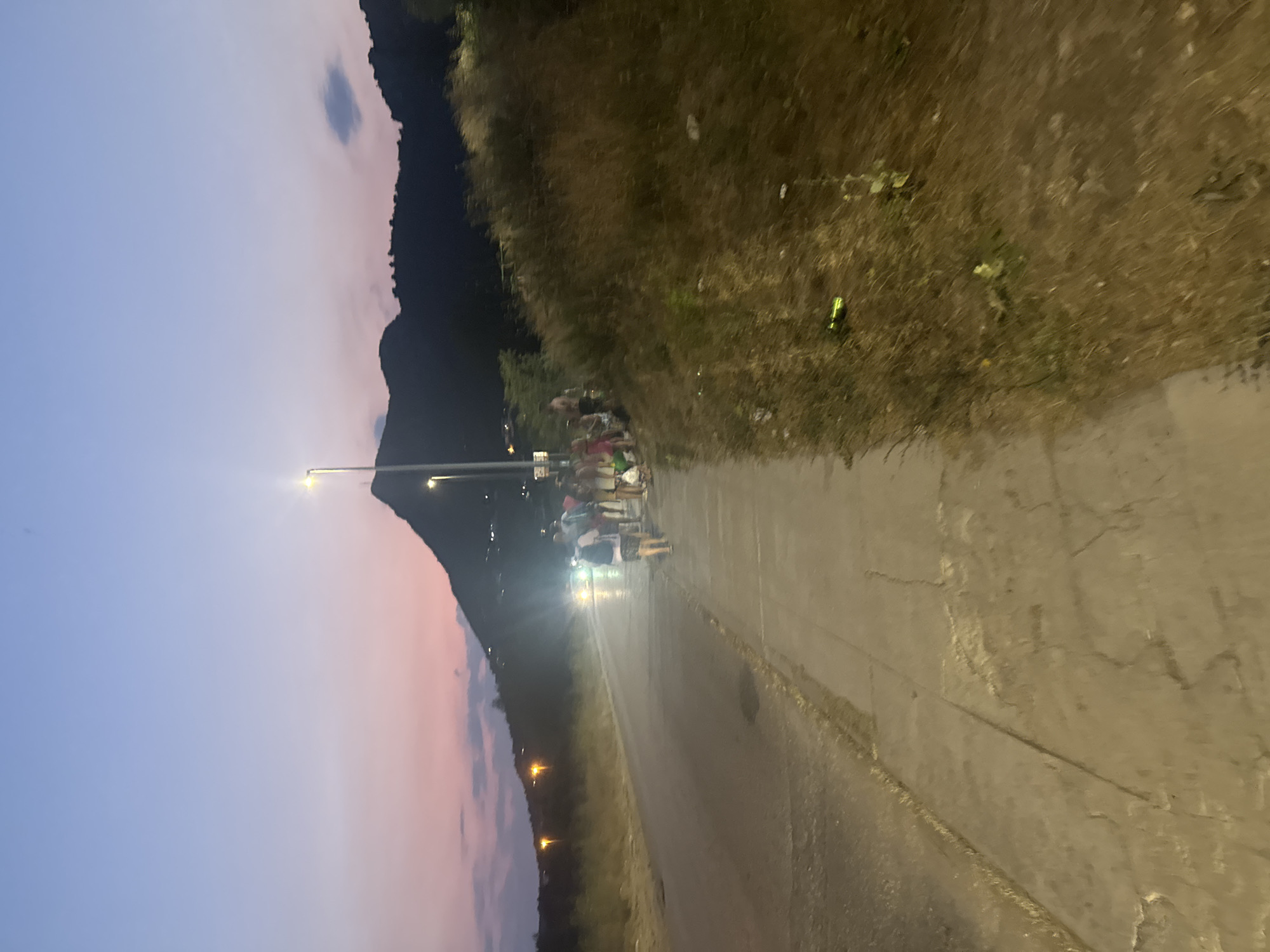 The image depicts a twilight scene along a paved road with a sidewalk in Sutomore, Montenegro. A group of approximately ten individuals, appearing to be tourists or locals in casual attire, are gathered on the sidewalk. Some are walking, while others are stationary, conversing. A car with headlights on is approaching on the road, its lights illuminating the scene. Beyond the road, dry brush and vegetation cover a sloping hillside. In the background, silhouetted mountains rise against a sky displaying hues of pink and purple, characteristic of a sunset or twilight. Streetlights are visible, casting artificial light on the sidewalk. The overall environment suggests a public area, possibly a coastal town, during a transition from day to night.