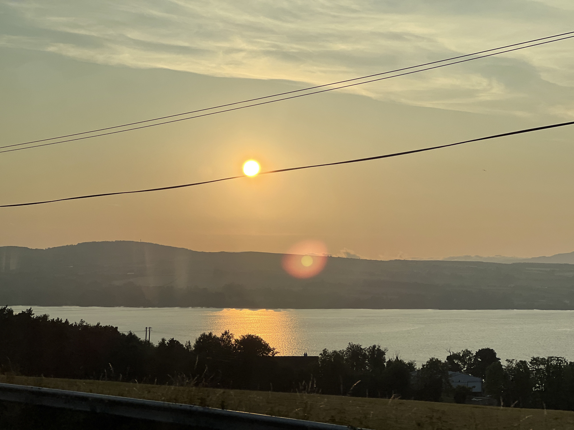 The image captures a serene sunset over a body of water, likely Lough Swilly, as viewed from an elevated position on the outskirts of Letterkenny, Ireland. The sun, a bright orange orb, is partially obscured by two horizontal power lines, casting a warm glow across the sky and water. Wispy clouds stretch across the upper portion of the frame. In the middle ground, a gentle slope of hills is visible, silhouetted against the fading light. The foreground is dominated by dark trees and foliage, with a glimpse of a road barrier. The overall impression is one of natural beauty and tranquility at the end of the day.