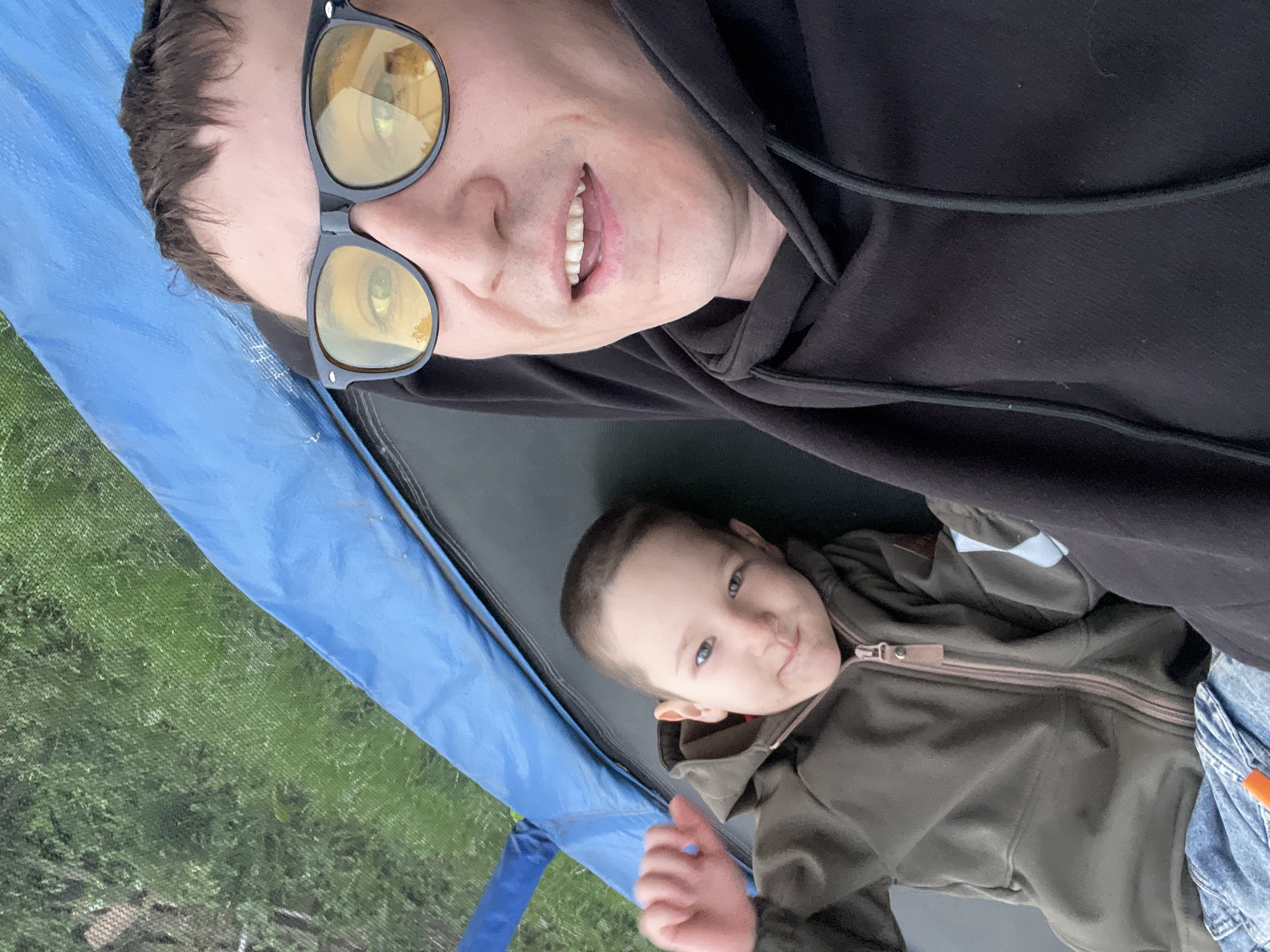 The image is a selfie taken from a high angle within what appears to be a trampoline enclosure. A man wearing black sunglasses and a black hoodie is in the foreground, looking at the camera and smiling. Beside him, a young child with short hair and a green hooded jacket is also looking at the camera. The background shows the green netting of the trampoline and some out-of-focus greenery, suggesting an outdoor setting, possibly in Laishevo, Russia. The blue material of the trampoline mat or enclosure is visible in the upper left quadrant of the image.