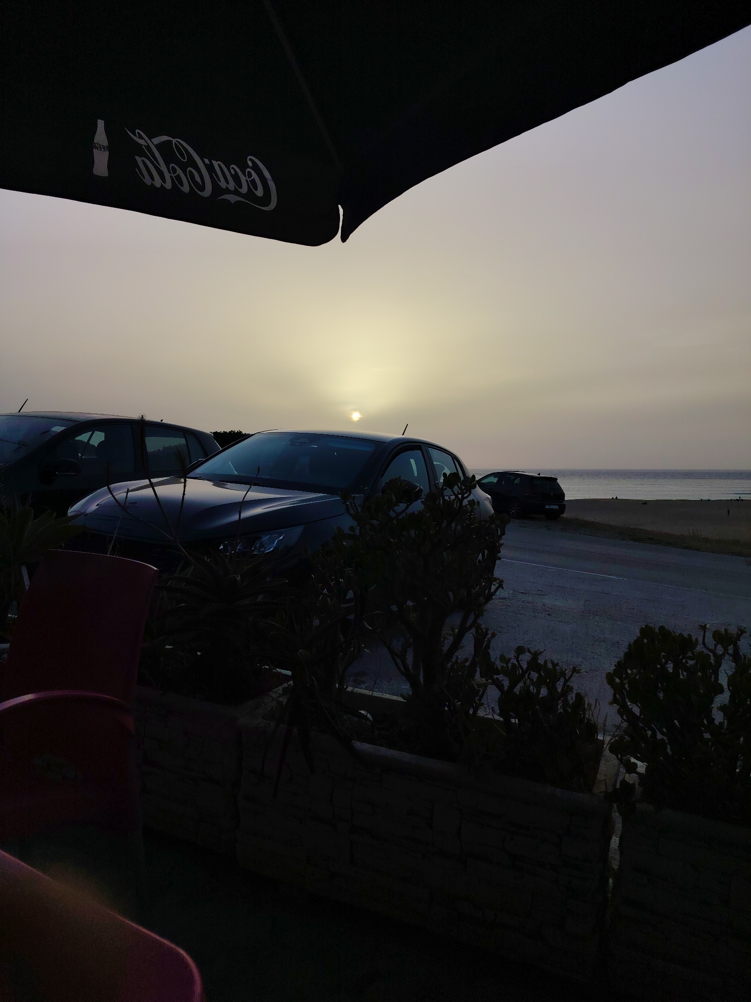 This image depicts a dusk scene near the coastline of Tangier, Morocco, possibly from a vantage point with outdoor seating. In the foreground, a dark-colored umbrella with a reflective Coca-Cola logo is visible, partially obscuring the sky. Several parked cars are present, with one dark sedan prominently positioned in the middle ground, its headlights off. Behind the cars, a calm sea stretches towards the horizon, with the sun setting low in the sky, casting a soft, diffused light. The immediate foreground also shows elements of outdoor furniture, including a red chair, and dense shrubbery. The overall atmosphere is serene and twilight-like, with the urban landscape of Tangier suggested by the presence of cars and infrastructure near the sea.