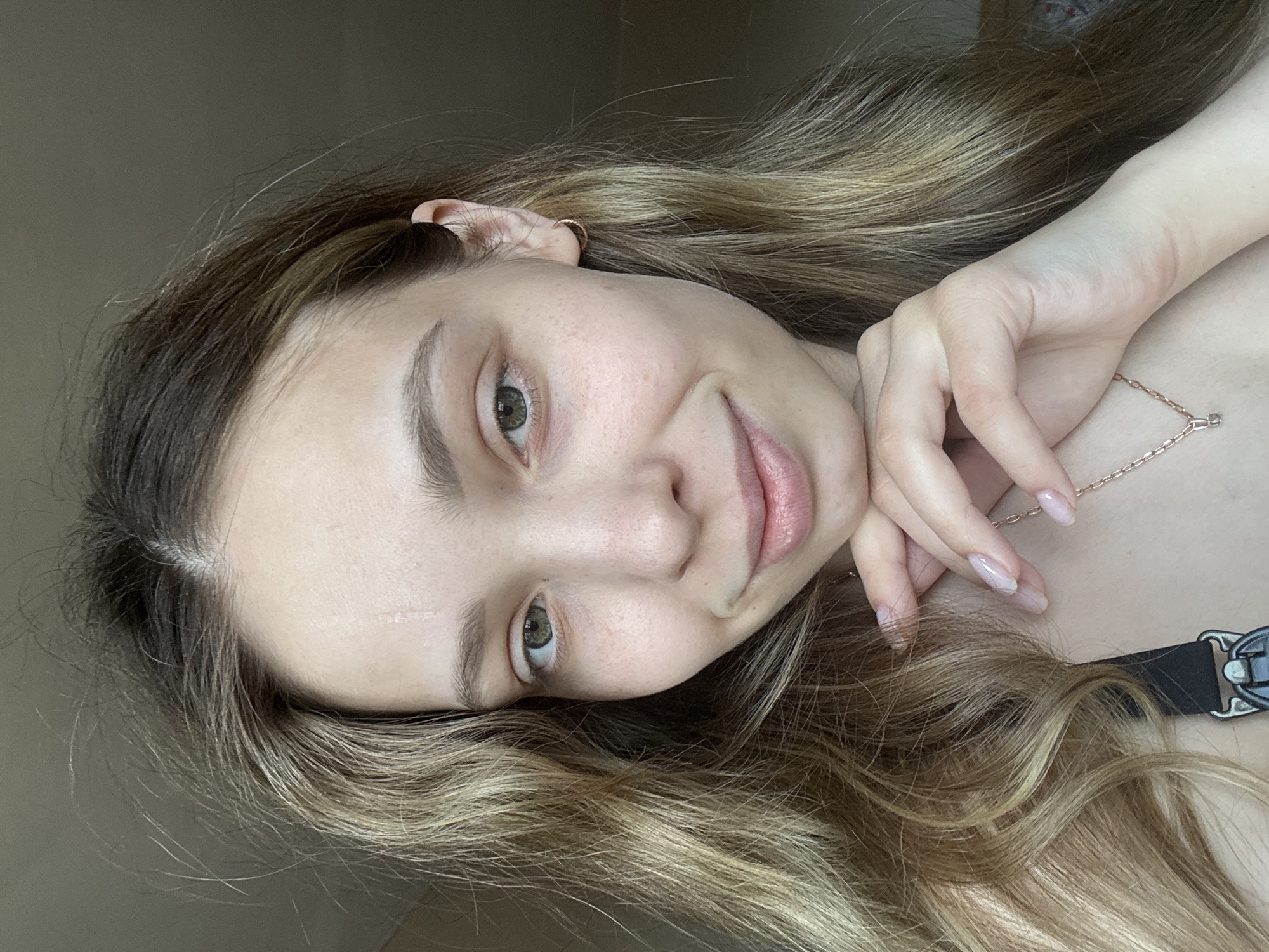 A young woman is photographed at an angle, capturing a close-up of her face and upper body. She has light skin, green eyes, and light brown hair with blonde highlights. A small gold hoop earring is visible in her left ear. She wears a delicate gold necklace. Her nails appear to be manicured. The background is a neutral beige color. The scene's location in the city of Chaikovsky, Russia is not discernable from the given image.