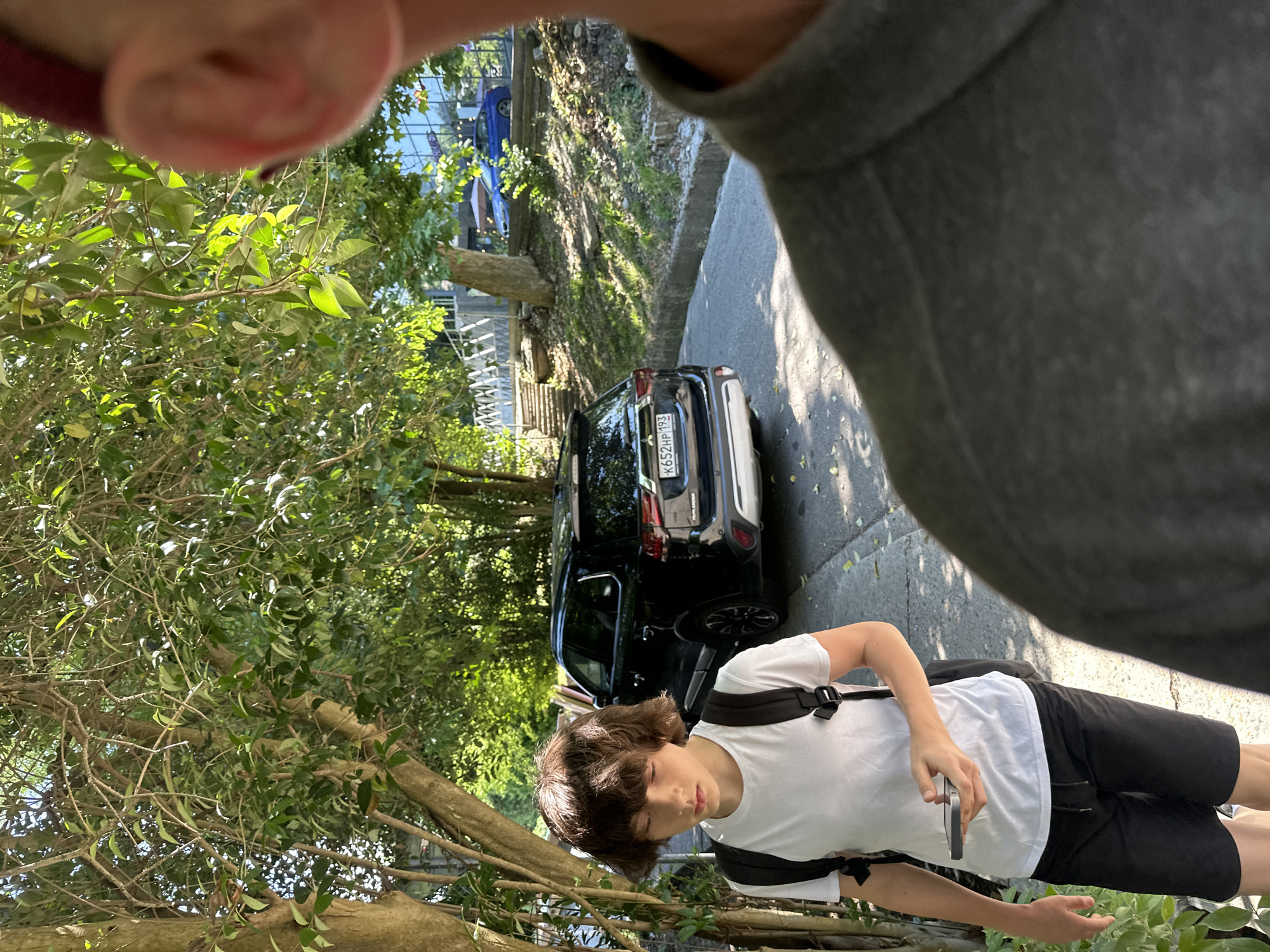 The image depicts a street scene in Sochi, Russia, on a sunny day. In the foreground, a person is captured from the chest up, facing away from the camera, with their head slightly turned. Their shoulder and neck are visible, clad in a dark gray garment. Further into the scene, a young person with brown, shaggy hair walks towards the left, looking down at a mobile phone held in their right hand. They are wearing a white t-shirt and black shorts, with a black backpack slung over their shoulders. A black SUV is parked on the right side of the road, with its license plate visible: K652HP 193. The background shows a leafy green environment with trees casting shadows on the asphalt road. Steps leading up a slope and some buildings are visible in the distance. The overall impression is of a casual outdoor setting.