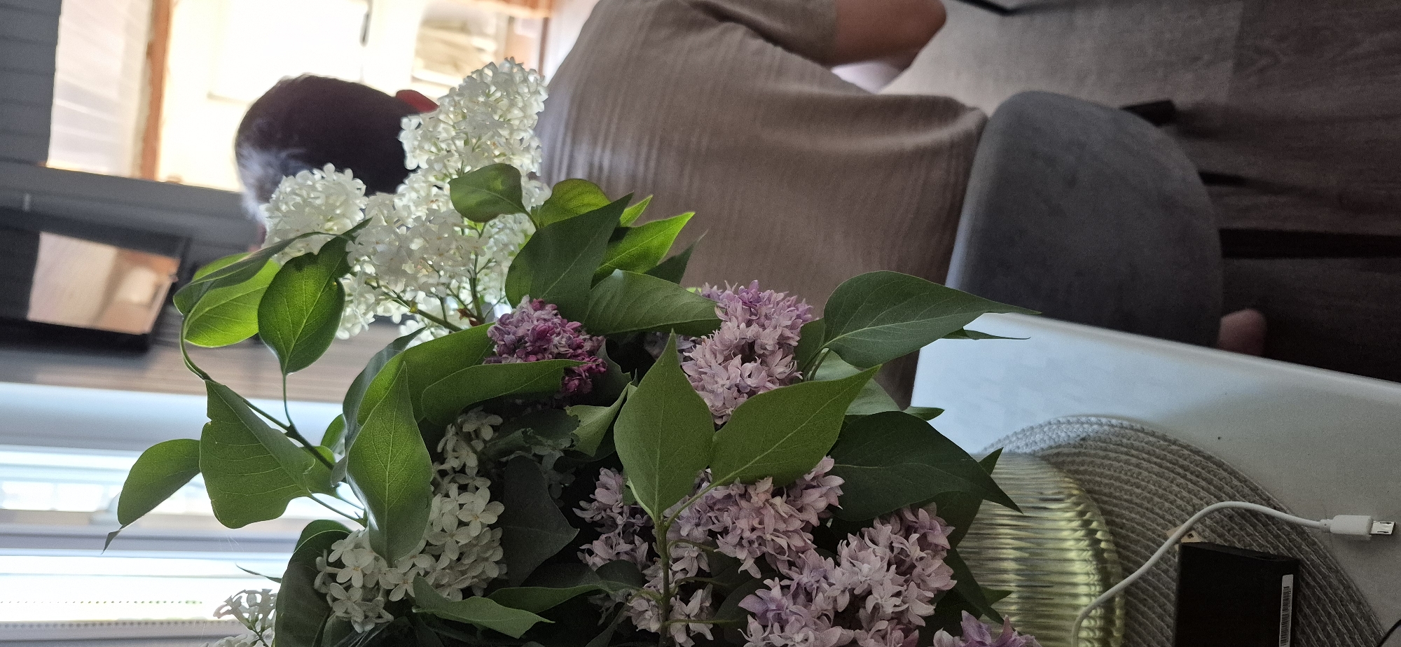 A bouquet of lilac flowers in shades of white and lavender sits on a table. The bouquet is composed of several clusters of flowers interspersed with green leaves. Behind the bouquet is a person wearing a beige shirt and seated on a grey chair. A white table surface is visible, along with a braided mat and a black electronic device connected to a white cable. The location is Unknown, Unknown.