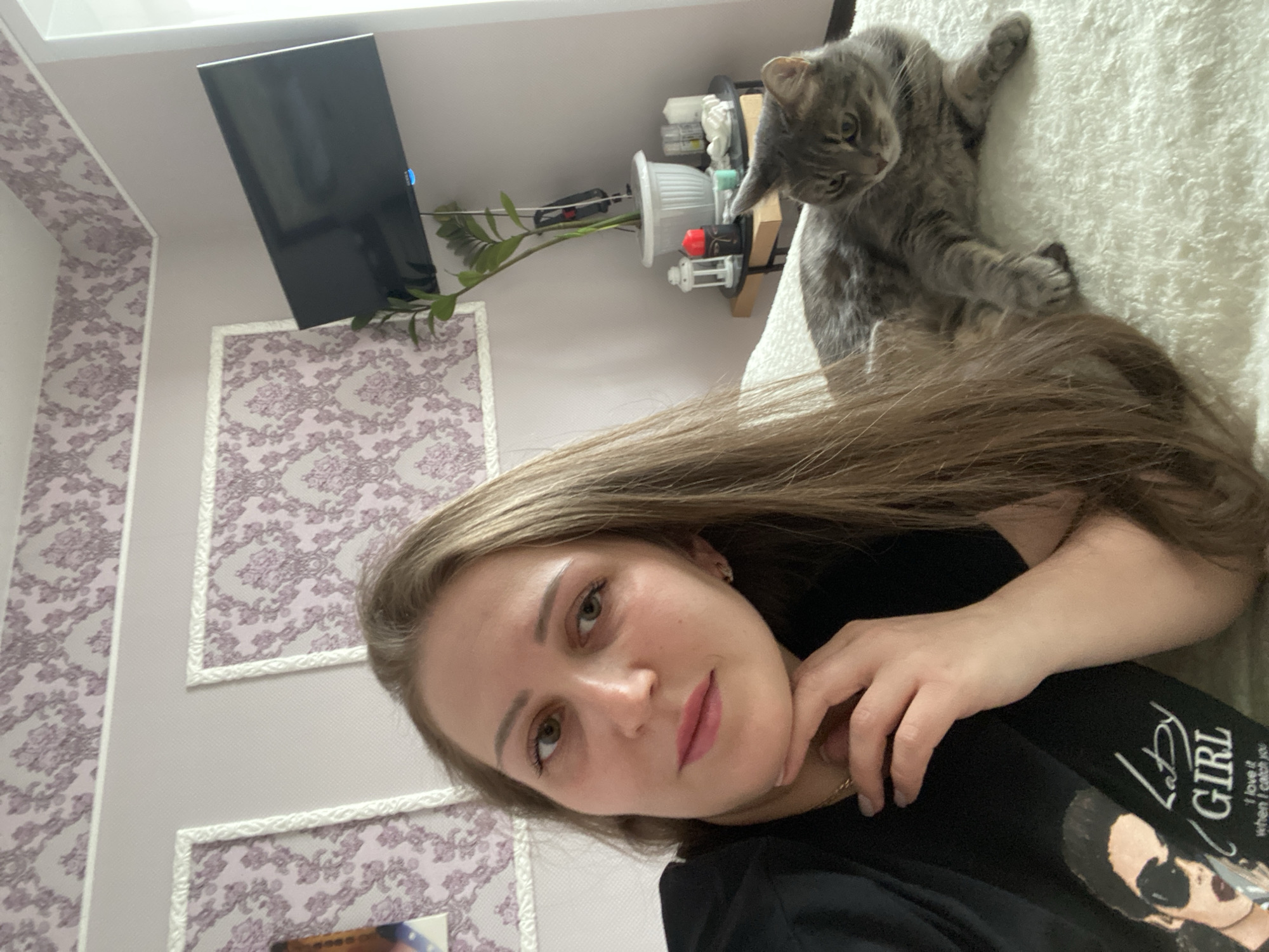 The image shows a woman lying down, with a gray tabby cat sitting on a light-colored, textured surface next to her. The woman has long blonde hair, fair skin, and is wearing a black shirt with the words "Lady Girl" visible. Her left hand is near her face. A TV is mounted on the wall in the background. A potted plant and shelf-like structure are adjacent to the TV. The wall has a patterned wallpaper. The location is Markovskii, Russia.