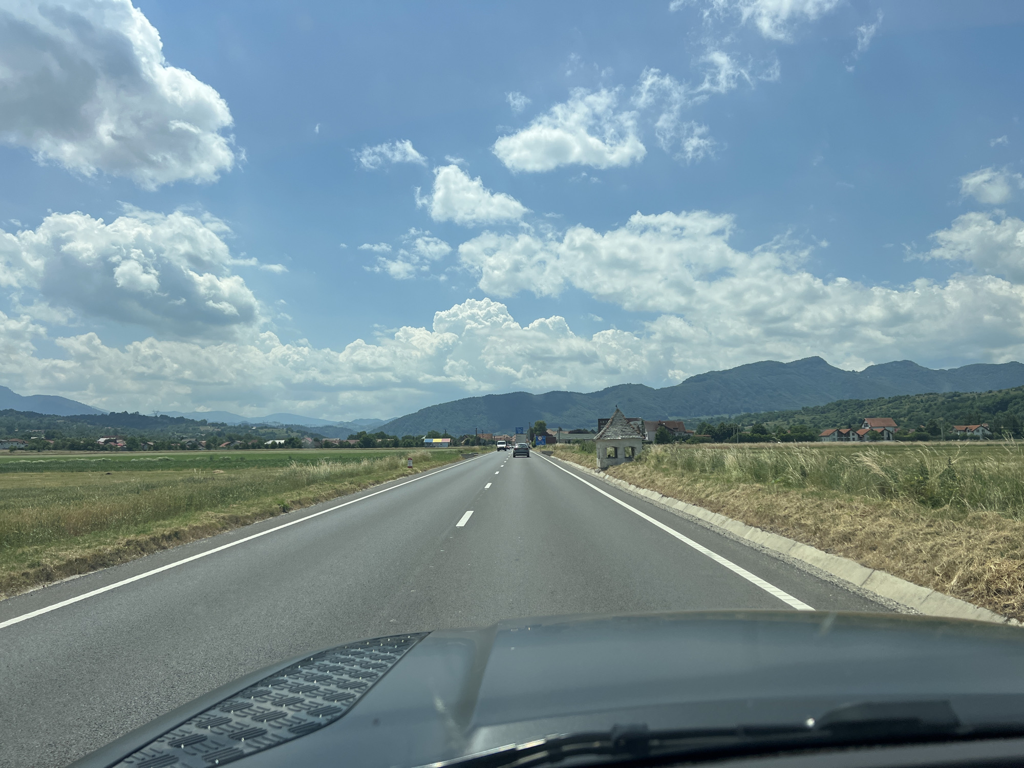 The image depicts a view from the front of a vehicle driving on a two-lane asphalt road. The road is bordered by grassy fields and a concrete curb. In the distance, there are rolling green hills and mountains. A few scattered houses and buildings are visible along the road and nestled in the foothills.
The sky is a vibrant blue, filled with numerous fluffy white clouds. The time of day appears to be daytime, possibly late morning or early afternoon, given the bright sunlight and the shadows cast. The weather seems pleasant and clear, with the sun illuminating the landscape.
There are a few cars visible on the road ahead, indicating that this is a public roadway in use. On the right side of the road, there is a small, old-looking structure that resembles a rustic shelter or a historical marker. The foreground shows the hood of the vehicle, with a textured surface, and the windshield wipers. The scene evokes a sense of travel and a journey through a rural or semi-rural landscape. There are no discernible people or explicit text in the image.