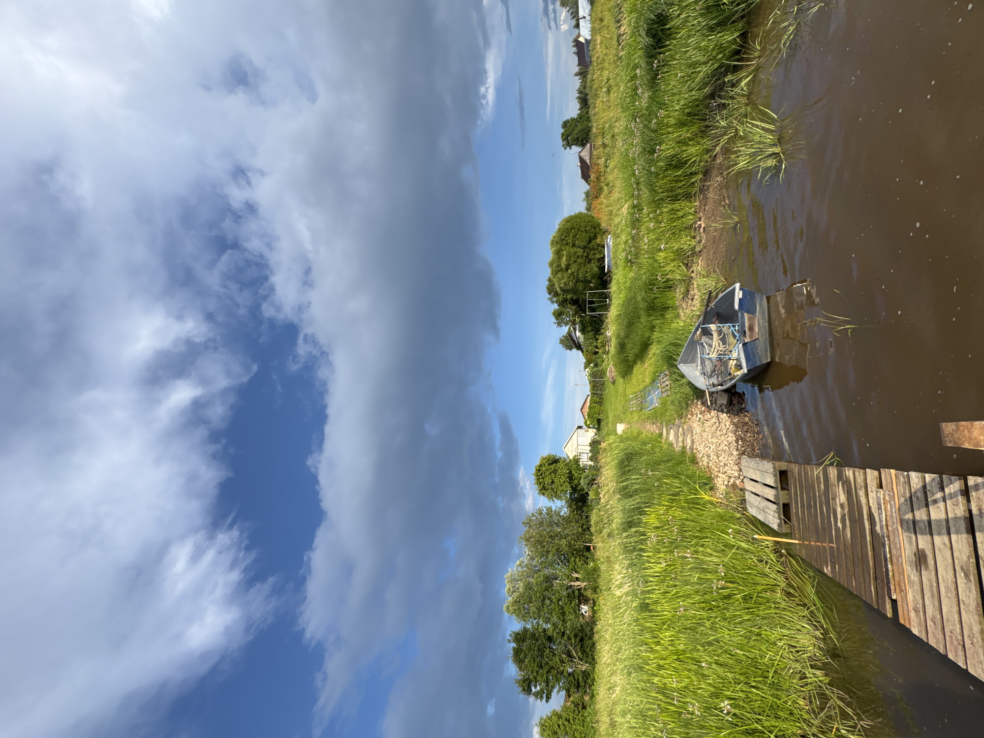 The image depicts a rural landscape in Sitno, Russia, on a cloudy day. In the foreground, a weathered wooden pier extends into murky brown water. A small, blue motorboat is moored alongside the pier, its interior containing fishing nets. The pier leads to a gravel path that winds through tall, lush green grass dotted with small white flowers. In the middle ground, several trees stand to the left, with a white house and other structures visible in the distance to the right, nestled amongst fields and more trees. The sky above is a dramatic mix of bright blue patches and dark, heavy clouds, suggesting an approaching storm. The overall impression is one of natural beauty and tranquility in a remote setting.