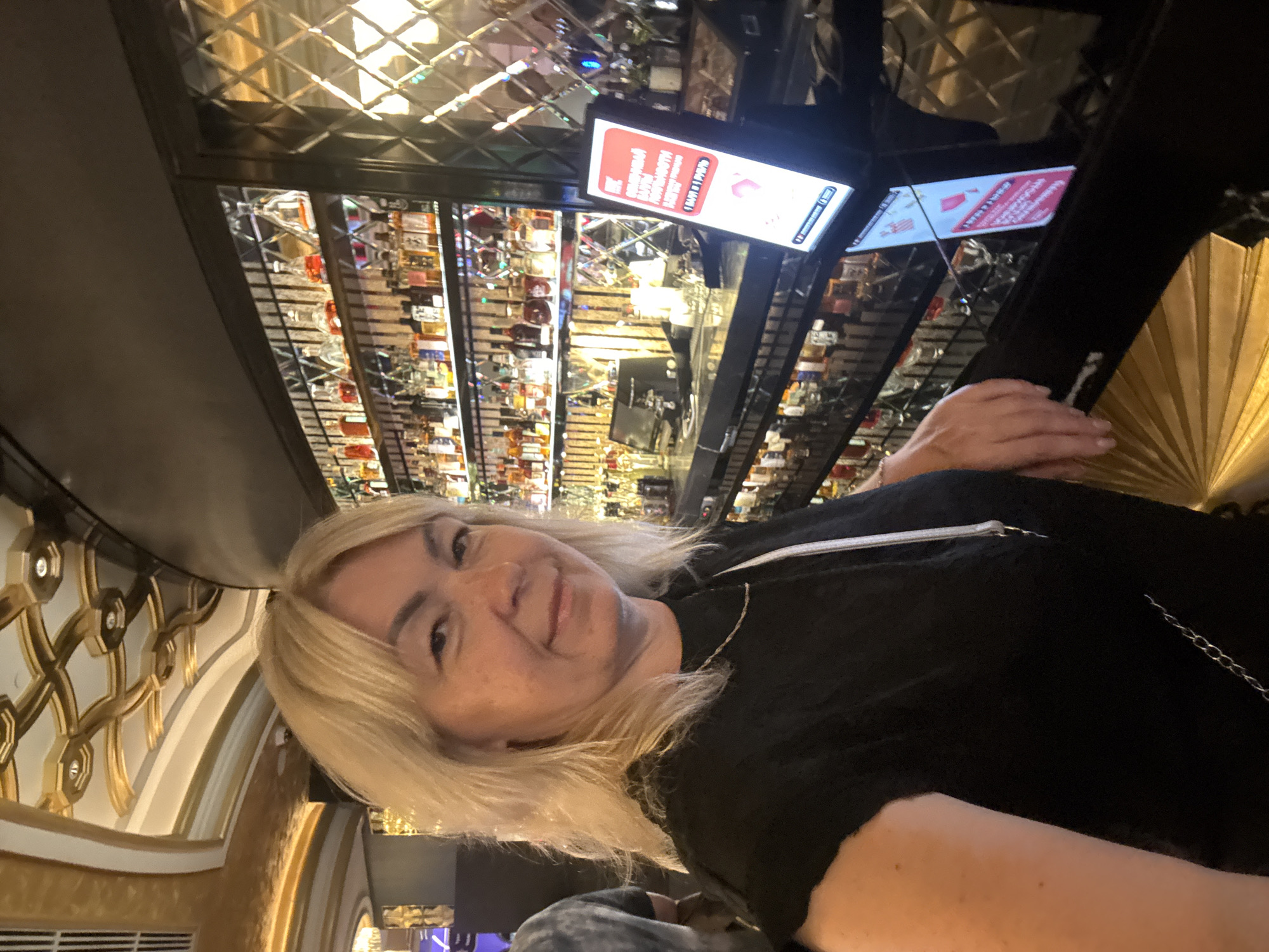 A woman with blonde hair is pictured in a dimly lit room, possibly a bar or lounge. She is wearing a black top and appears to be smiling. Behind her, shelves are filled with numerous bottles, suggesting a liquor display. The background features decorative elements, including a patterned ceiling and ornate gold trim on the wall. Two screens are visible in the background, one displaying text in red and white. The overall ambiance suggests a social gathering or evening outing. The image is oriented sideways.