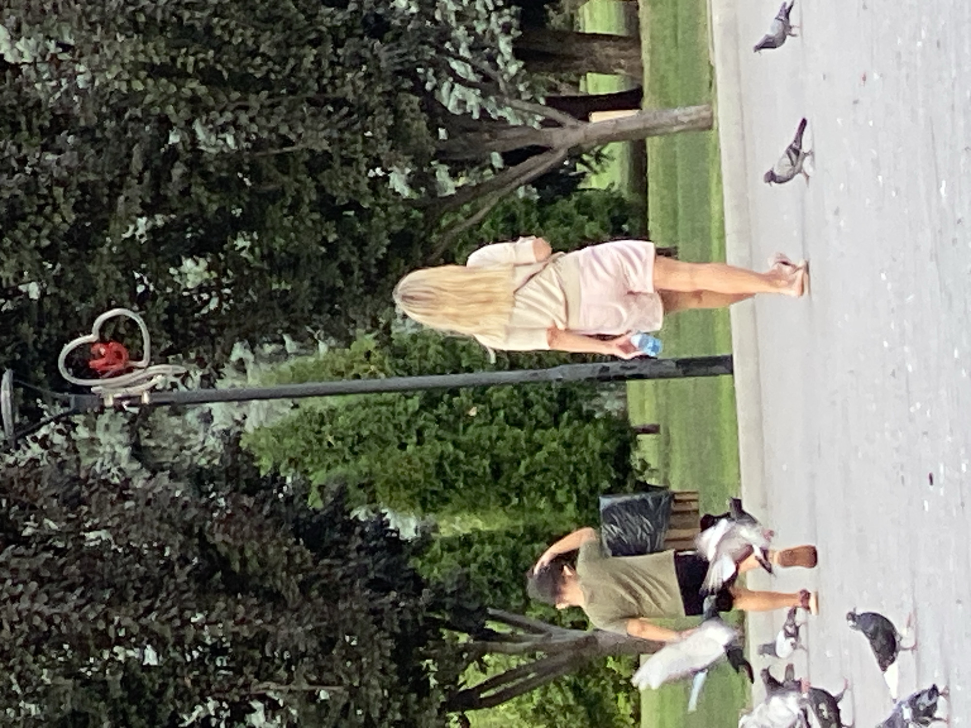 In a paved outdoor area, possibly a park or promenade, a young boy and an adult female are walking away from the camera, surrounded by pigeons. The boy, on the left, is wearing a dark green t-shirt and dark shorts. He has his left hand on the back of his head and is holding something white in his left hand, possibly a toy. The woman, on the right, has long blonde hair and is wearing a light-colored t-shirt and light pink shorts. She is holding a blue water bottle in her right hand. Several pigeons are visible on the ground around them, with some in mid-flight, suggesting the people may be interacting with or feeding them. In the background, lush green trees and a lamppost with a decorative heart shape are visible. The scene is set in Chișinău, Moldova.