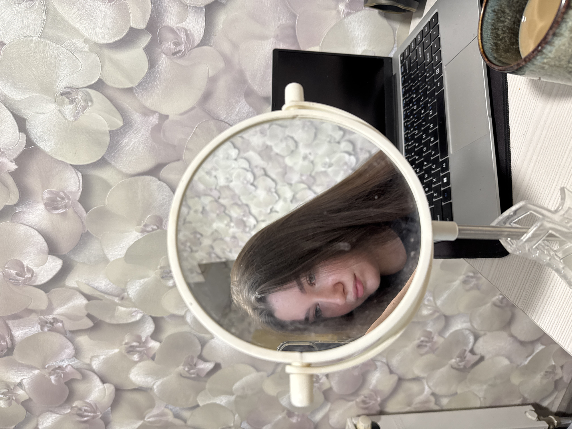 A top-down view depicts a round, white-framed vanity mirror reflecting a young woman with long, dark brown hair and fair skin. Her head is tilted, and she gazes into the mirror. The mirror is positioned on a table with a light-colored, floral-patterned wallpaper visible in the background. A laptop and a mug of liquid are partially visible to the right of the mirror, with a plastic object adjacent to the laptop. No specific details confirm the location as Perm, Russia.