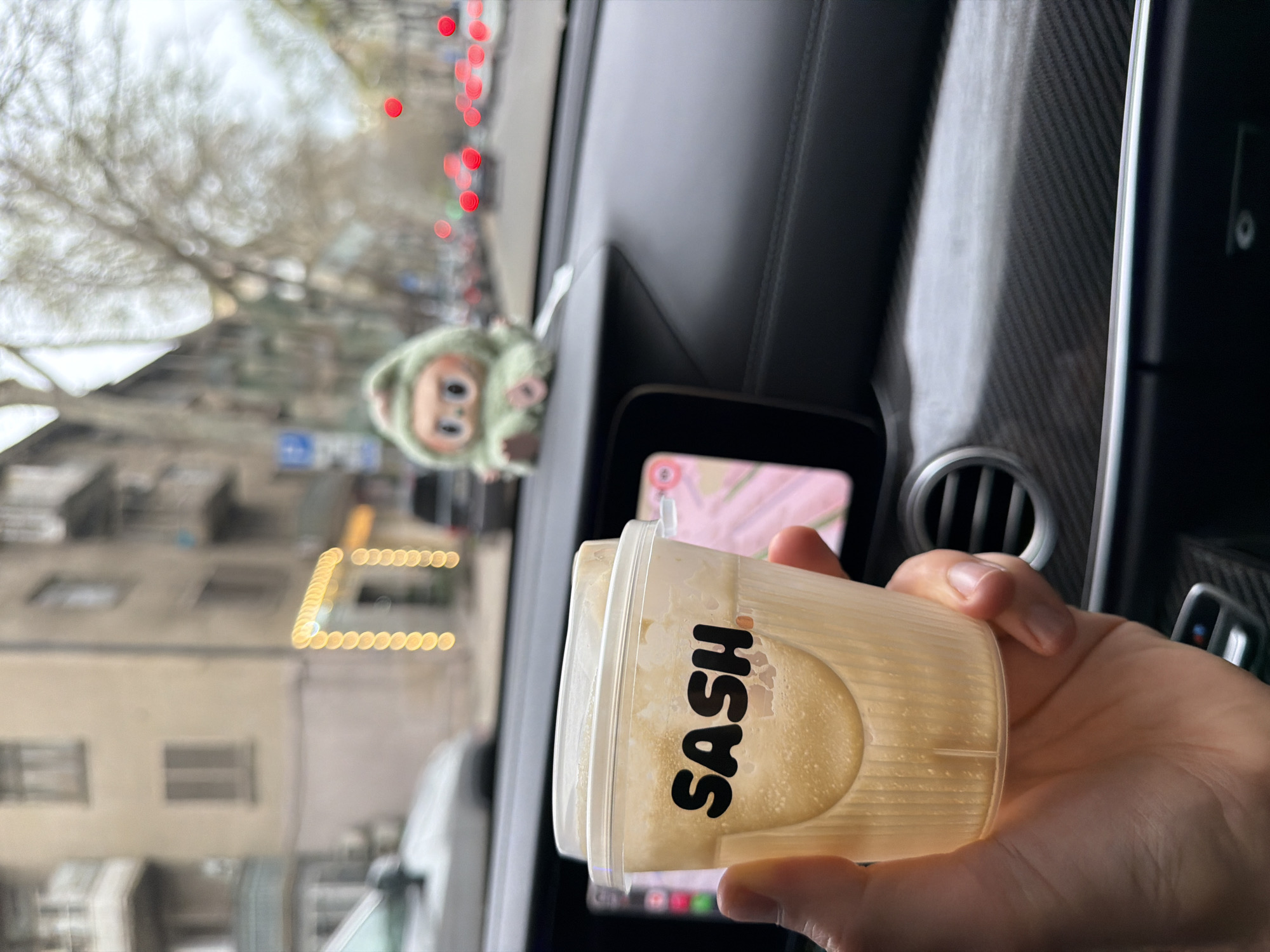 A person inside a car is holding a clear plastic cup with the word "SASH" printed on it in black, filled with a light-colored liquid, likely a beverage. The car's interior includes a textured dashboard, air vents, and a touchscreen displaying a map. A small, green plush toy hangs from the rearview mirror. The outside environment shows a city street with buildings and trees, blurry lights, and a building with yellow decorative lights. Based on the architectural style, the environment, and the cup with "SASH," the location is likely Tbilisi, Georgia.