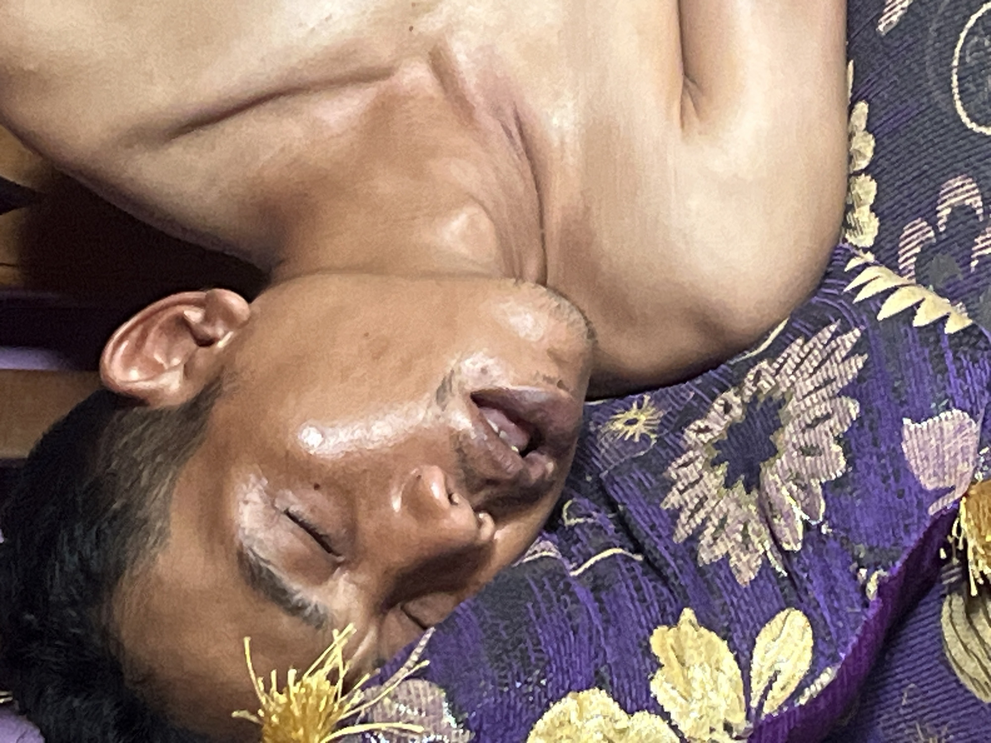 A close-up, over-the-shoulder shot captures a person lying on their side on a purple, floral-patterned fabric, possibly a sarong or bedspread. The person's face, partially obscured by their hair, is turned downwards, with eyes closed and mouth slightly open, suggesting sleep or unconsciousness. A small, golden, tassel-like object is positioned near their left eye. The person appears to be unclothed from the chest up. The lighting in the image is dim, highlighting the contours of the person's face and shoulder. The scene is set indoors, with a wooden surface visible in the background, hinting at a domestic environment in Karang Sari, Indonesia.