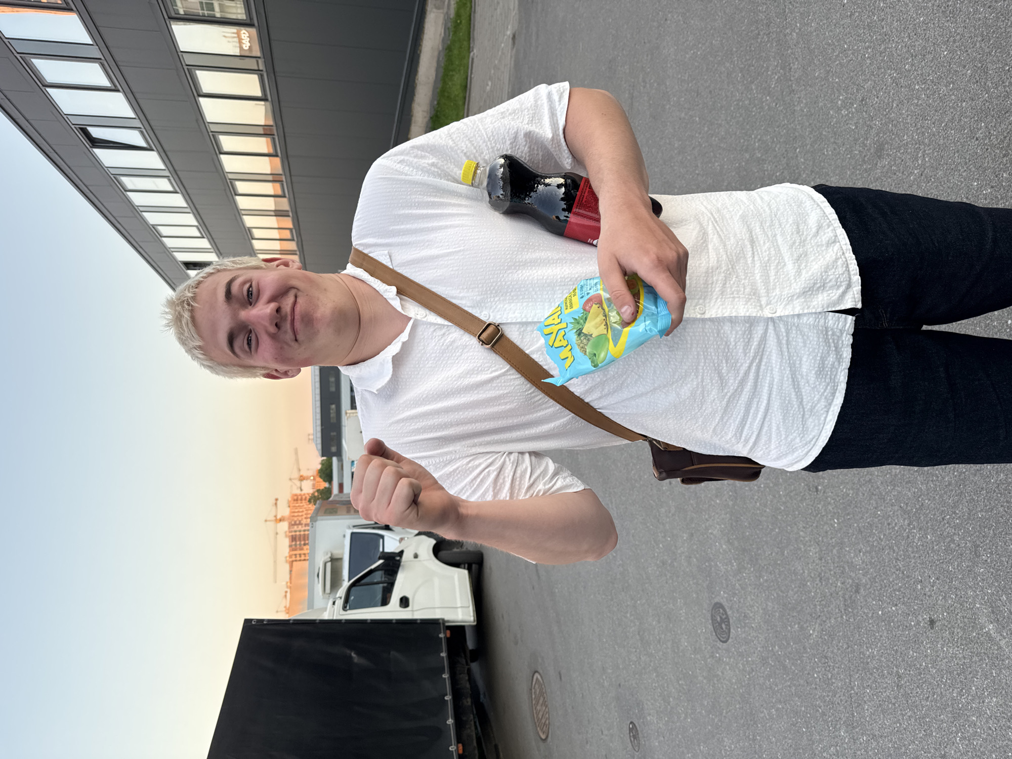 A young male with short, bleached blonde hair, a light complexion, and a faint red mark on his left cheek stands in an outdoor asphalt area, smiling broadly at the camera. He wears a white textured short-sleeved button-up shirt, dark jeans, and a brown leather cross-body bag with the strap visible over his right shoulder. His right hand is clenched in a fist, held slightly upwards. In his left arm, he holds a light blue package of "MAYA" branded fruit snacks and a dark brown plastic bottle with a yellow cap and a red label. To his left, a large black truck is parked next to a white delivery van. Further in the background, multiple construction cranes and partially built structures are visible against a light orange and blue sunset sky. To his right, a modern dark grey building with multiple reflective rectangular windows, one of which is open, stands next to a patch of green grass. The ground is grey asphalt with a circular manhole cover. The scene is located in the city of Unknown, Unknown.