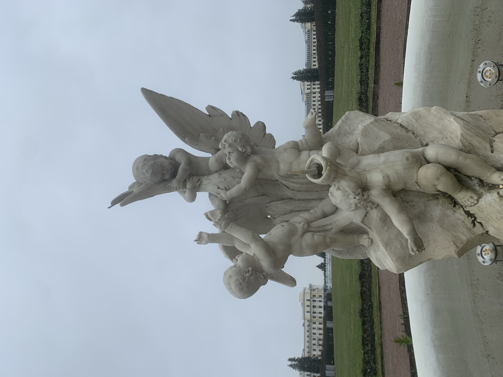 A close-up, slightly angled shot shows a white stone sculpture featuring several cherubs or putti figures, some with wings. The sculpture appears to be part of a fountain or similar decorative structure. Behind the sculpture is a grassy area, a reddish-brown pathway, and a large, cream-colored building with multiple stories and windows. The sky is overcast and gray. The scene is located in Arkhangel'skoe, Russia.