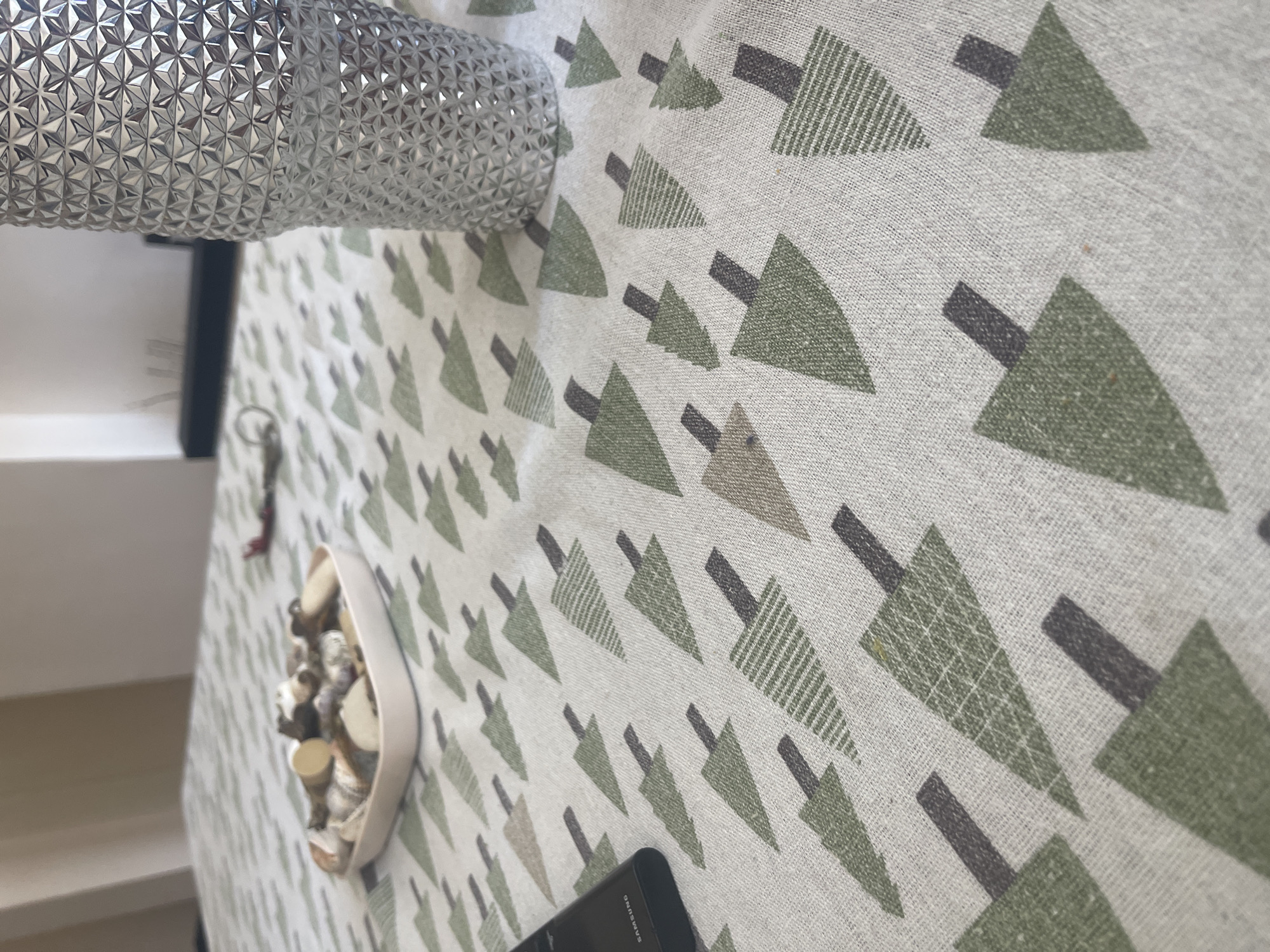 The image depicts a close-up, eye-level view of a table covered with a light-colored tablecloth featuring a repeating pattern of green and brown arrow-like shapes. To the right, a textured silver cup or vase is prominently positioned. In the middle left, a small, light-colored rectangular dish contains an assortment of small, light-brown and white objects, possibly decorative stones or shells. A dark-colored smartphone with the "SAMSUNG" logo is visible on the left edge of the frame. The background shows a blurred interior setting with hints of furniture and wall. There is no discernible information within the image to place it in Varna, Bulgaria, or any specific city.