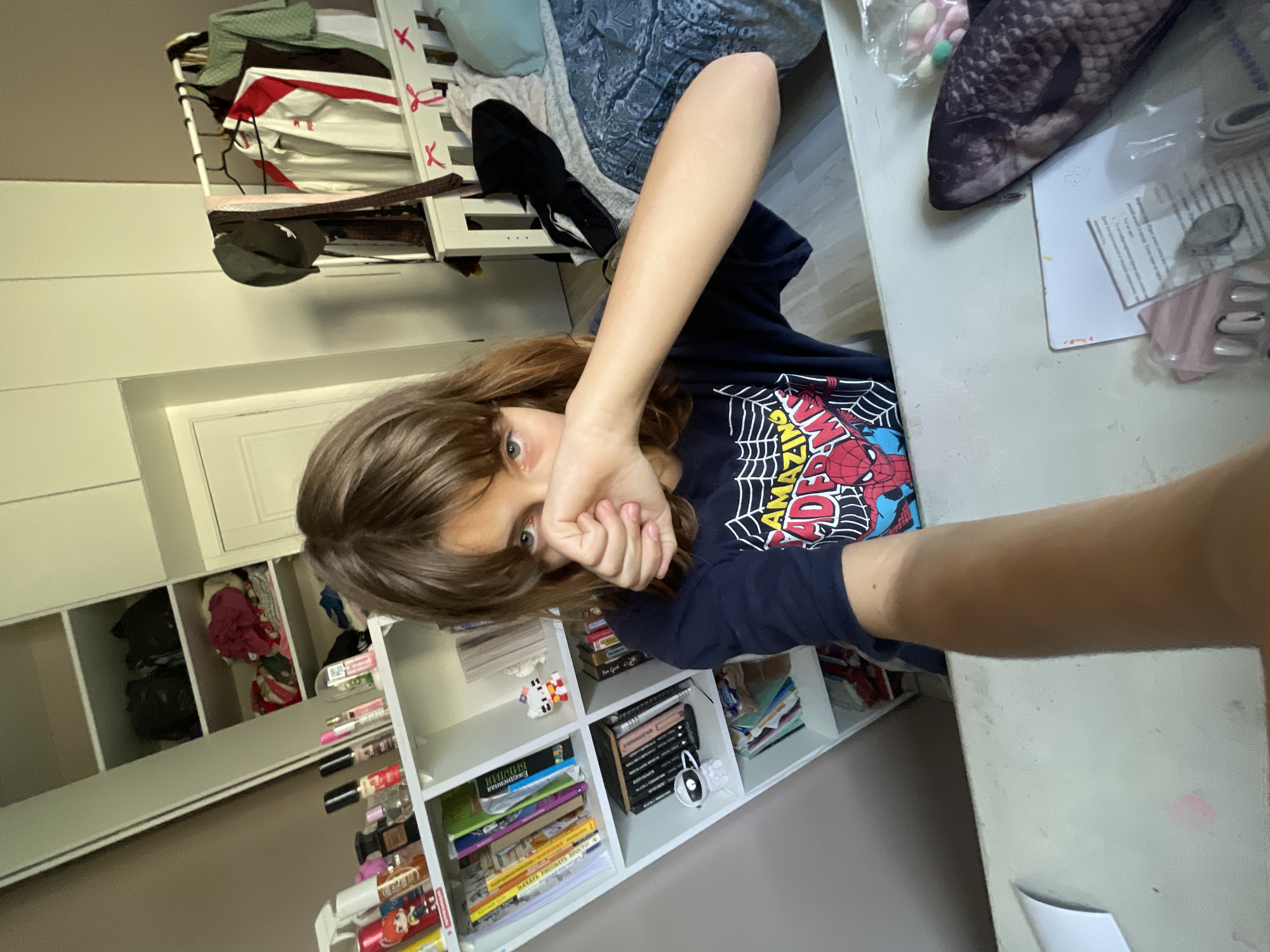 The image is a selfie taken indoors, likely in a bedroom. A young person with brown hair and bangs is looking at the camera, covering their mouth with their hand. They are wearing a dark blue t-shirt with a Spider-Man graphic. To the left of the person is a white bookshelf filled with books and various items, including what appear to be cosmetics and small figurines. To the right, a bed is visible with blue and grey patterned bedding, a headboard with decorative ribbons, and clothes hanging on a rack nearby. A dresser or wardrobe is also partially visible in the background. The lighting suggests it is daytime. The overall impression is a casual, personal space. There is no indication of the specific location within Мухавецкі сельскі Савет, Belarus, beyond it being an indoor residential setting.