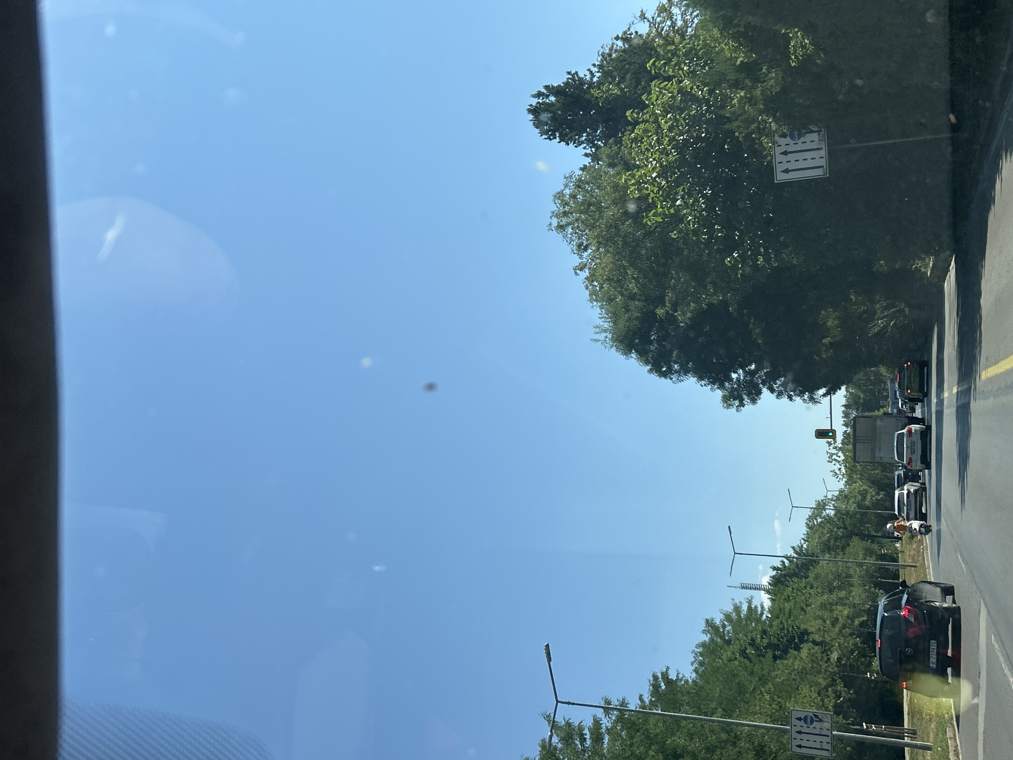 The image depicts a street scene in Sofia, Bulgaria, on a clear, sunny day. The perspective is from inside a vehicle, looking forward through the windshield. The road ahead has multiple lanes of traffic. Several cars are visible, including a black Opel Astra in the left lane, a white Audi, and a white SUV. A person on a scooter is riding between the lanes. On the left side of the road, there are trees and a streetlamp. A traffic sign indicating multiple lanes is also present. On the right side of the road, large trees line the street. Further down the road, a traffic light is visible, showing a green signal. The sky is a clear blue. The scene suggests a typical urban environment with moderate traffic.