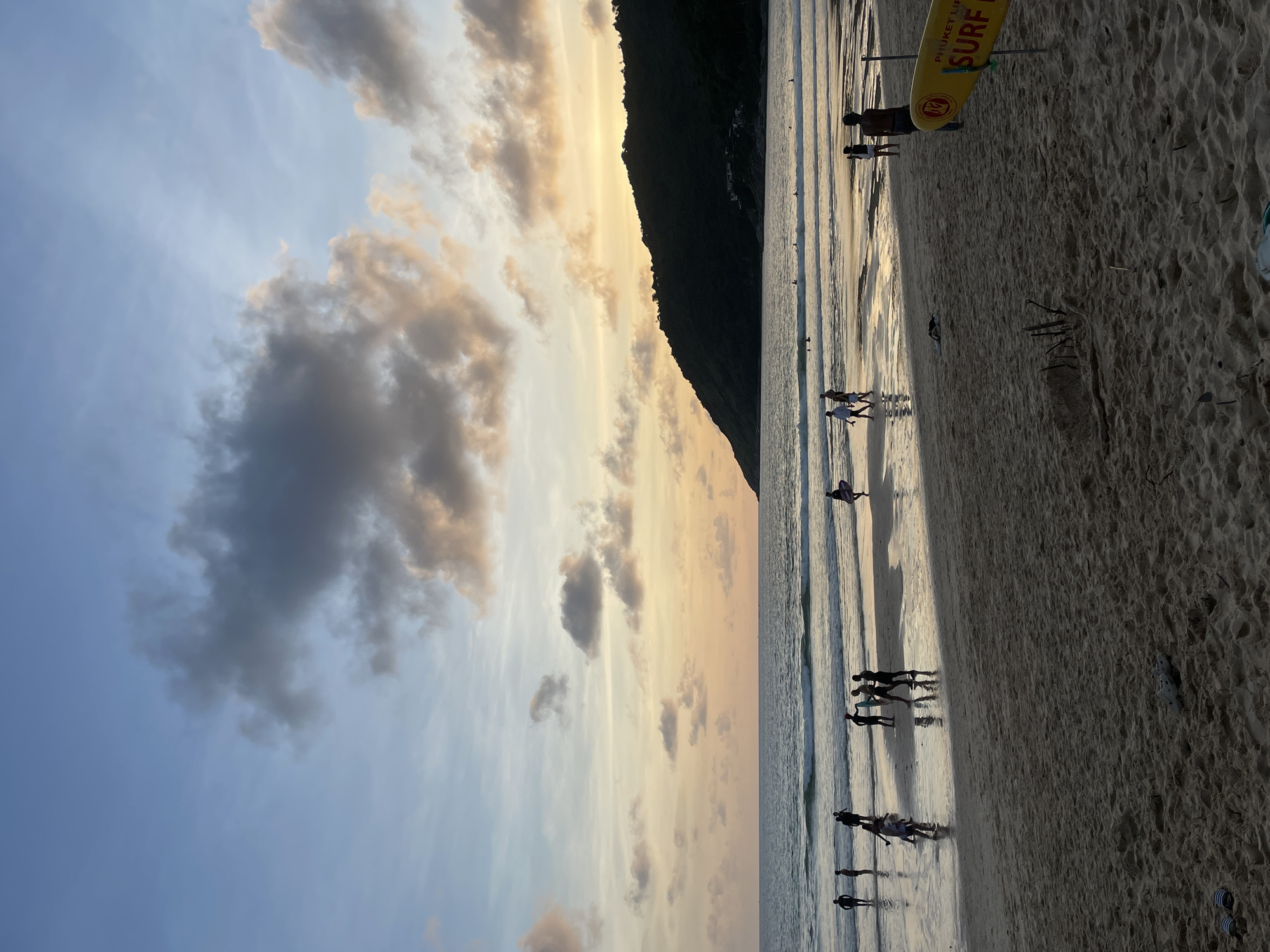 The image depicts a beach scene in Rawai, Thailand, during what appears to be sunset or dusk. The sky is filled with a mix of pale blue and warm hues of orange and yellow, with scattered clouds that are both dark and light. A dark, tree-covered hill or mountain dominates the right side of the horizon, with a few buildings visible on its lower slopes.
The sea is relatively calm, with gentle waves breaking on the shore. The wet sand reflects the colors of the sky and the surrounding landscape. Several groups of people are visible on the beach and in the shallow water. Some individuals are standing or walking, while others are gathered in small groups. A few people are holding surfboards.
In the foreground, the sandy beach is textured and shows some debris, including sticks and leaves. A yellow surfboard with red lettering that says "PHUKET LIFEGUARD SURF" is prominently displayed on a stand to the right of the image. Two pairs of sandals or flip-flops are visible on the sand in the lower left corner. The overall atmosphere suggests a peaceful evening at a popular tourist destination.