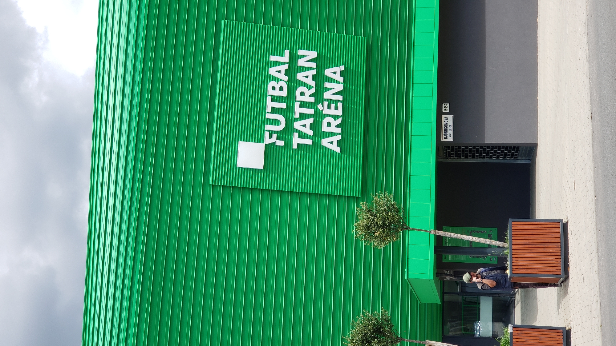 The image shows the Futbal Tatran Aréna in Prešov, Slovakia. The stadium has a distinctive bright green corrugated metal exterior. A white square logo is affixed to the green facade, with white text on it that reads "FUTBAL TATRAN ARÉNA". In the foreground, a man wearing a green cap and a dark vest over a t-shirt is standing next to a large wooden planter and talking on a mobile phone. Two small, potted trees are visible to the left and right of the man. A street sign with the text "BJÖRNSONOVA ULICA" and the number "16059" is mounted on the dark gray wall to the right of the entrance. The sky is cloudy.
