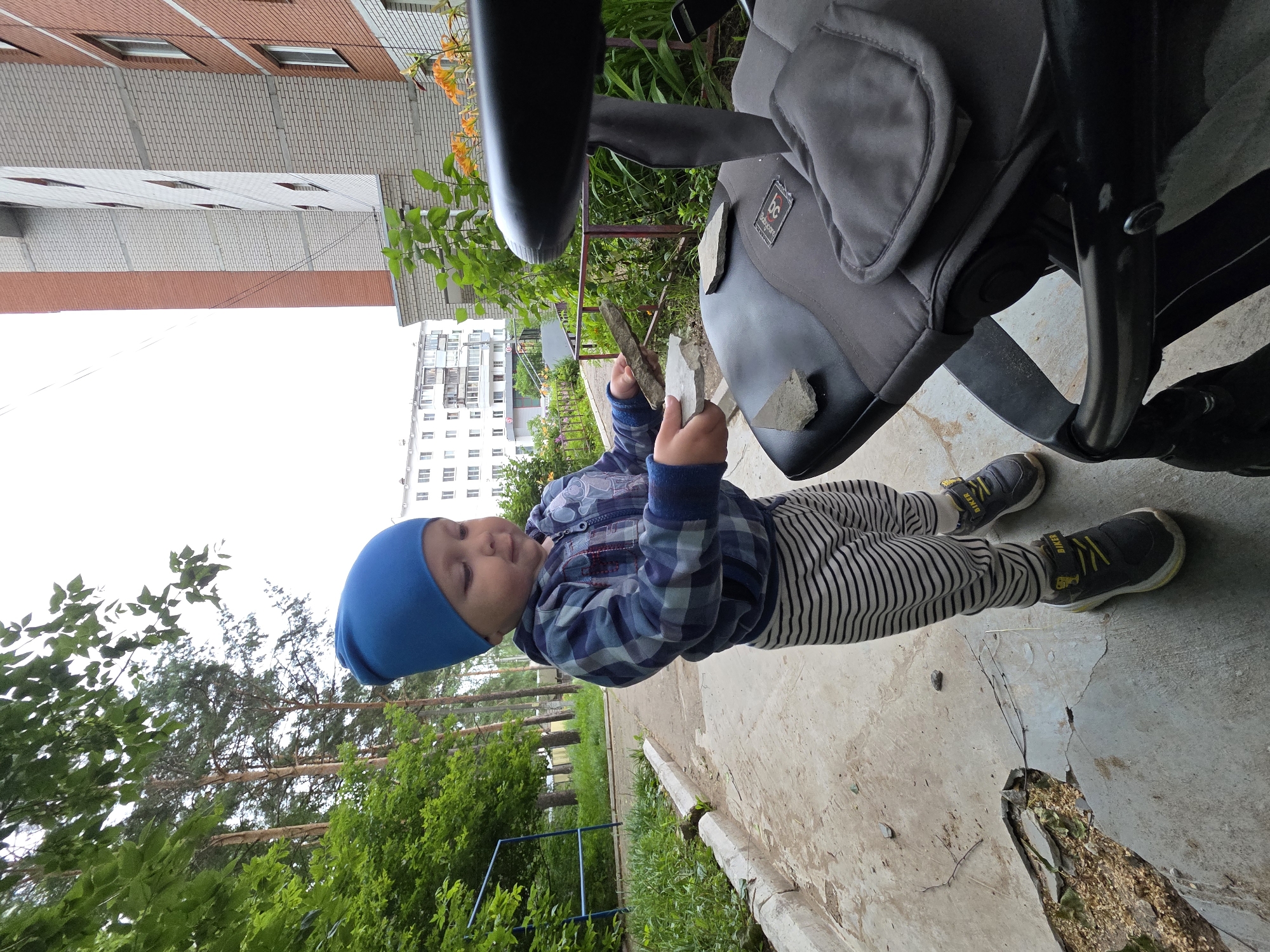 A young child wearing a blue beanie, a plaid jacket, and striped pants is standing on a concrete pathway next to a stroller. The child is holding two flat stones and looking down at them. The environment appears to be an outdoor area with trees, grass, and apartment buildings in the background. The stroller is dark gray and black. There are several other flat stones on the stroller's seat. The scene is likely in an urban residential area in Chaikovsky, Russia, given the architecture of the buildings.