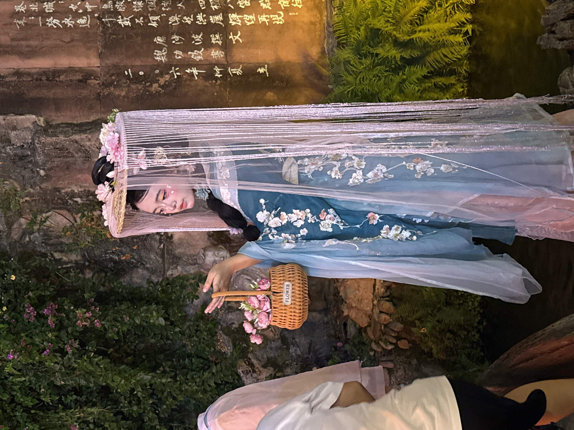 In Yuzhong District, China, a young woman in a traditional blue and white floral embroidered Hanfu dress stands adorned with a tall, veiled headdress featuring pink flowers and dangling beads. She holds a small woven basket filled with pink flowers, with a label reading "FLOWER" visible on the front. Her makeup includes subtle pink blush and artistic eye accents. Behind her, a stone wall is covered in green ivy and inscribed with Chinese calligraphy. To her left, another person is partially visible, wearing a light pink garment. The scene is set against a backdrop of lush green foliage, possibly a garden or park area, with a body of water or pond in the background.