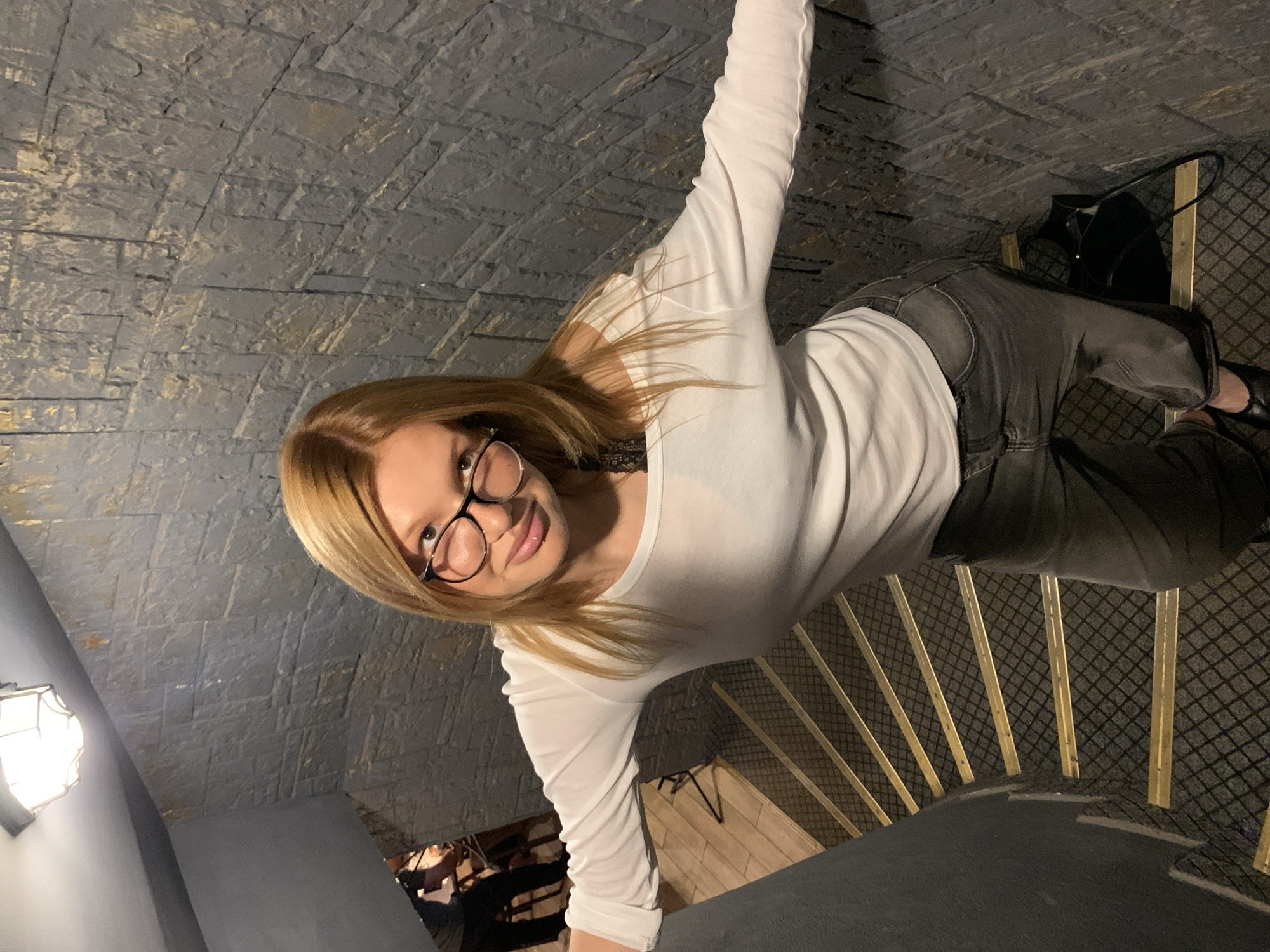 A woman with blonde hair and glasses is standing on a spiral staircase. She is wearing a white long-sleeved shirt and grey jeans. She is looking down at the camera with a slight smile. The staircase is made of stone and has wooden steps. There is a black bag on the stairs behind her. A light fixture is visible on the wall to the left. The scene appears to be indoors, possibly in a historic building. The time of day and weather are not discernible from the image. There is no visible text in the image.