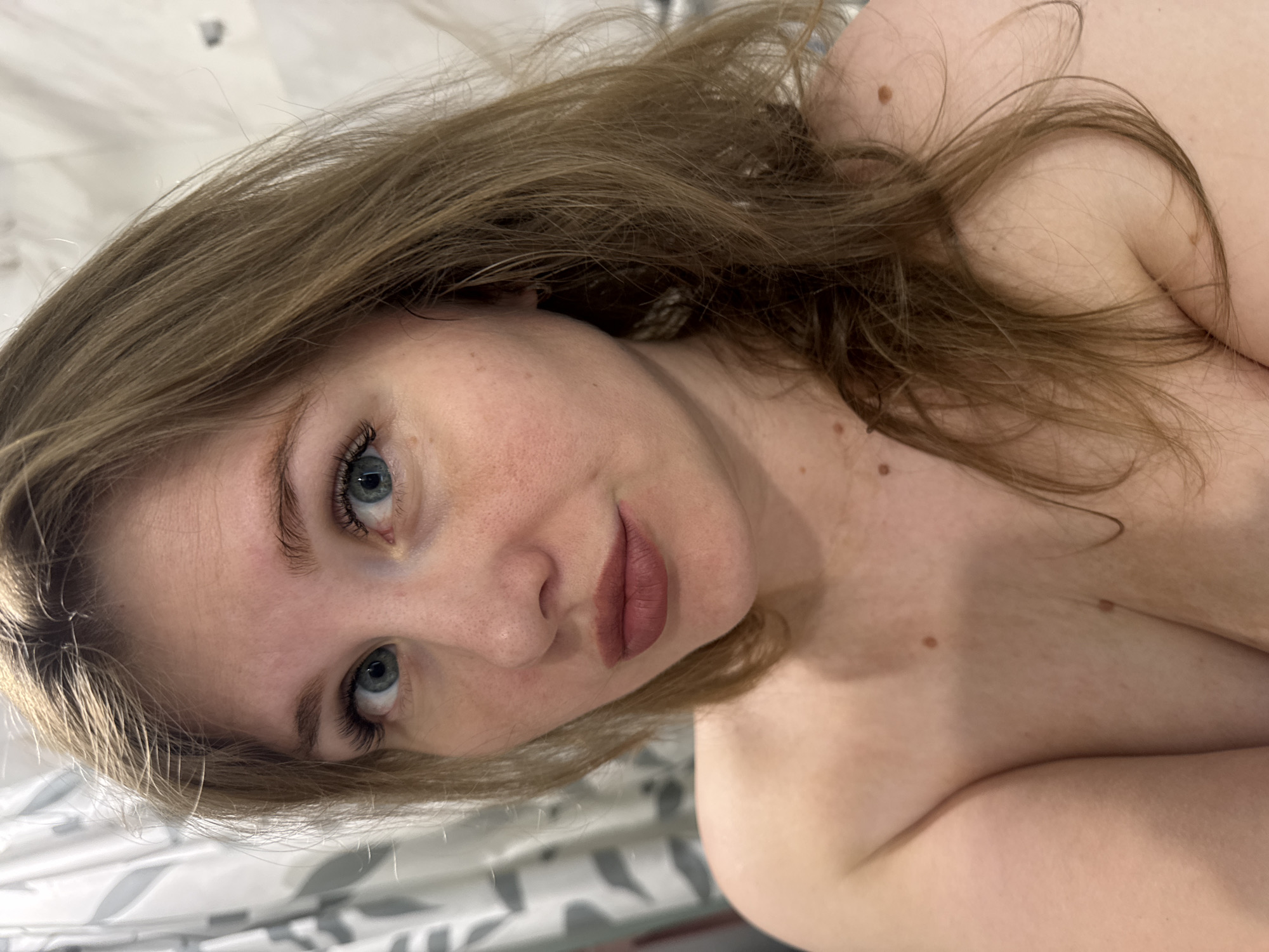 A close-up portrait depicts a fair-skinned woman with blue eyes and light brown hair. The camera angle is slightly tilted, with the woman looking directly at the viewer. Her facial features are prominent, including her eyelashes and lips. Her right shoulder and collarbone are visible, suggesting the presence of a torso. The background appears blurry, with a patterned object visible behind her. The image does not provide information about the city of Ферма, Russia.